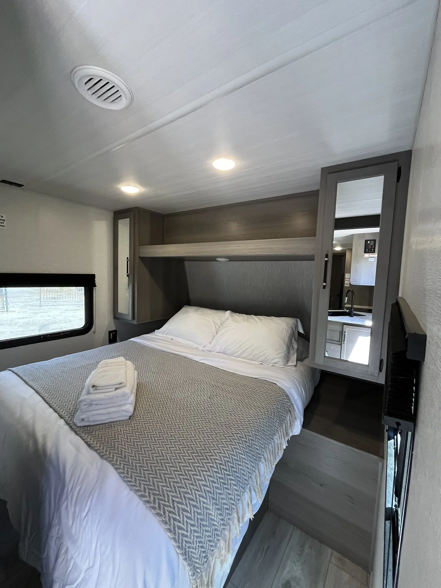 Interior of a bedroom in a recreational vehicle or camper with a bed, window, cabinet, overhead light, and a mirror.