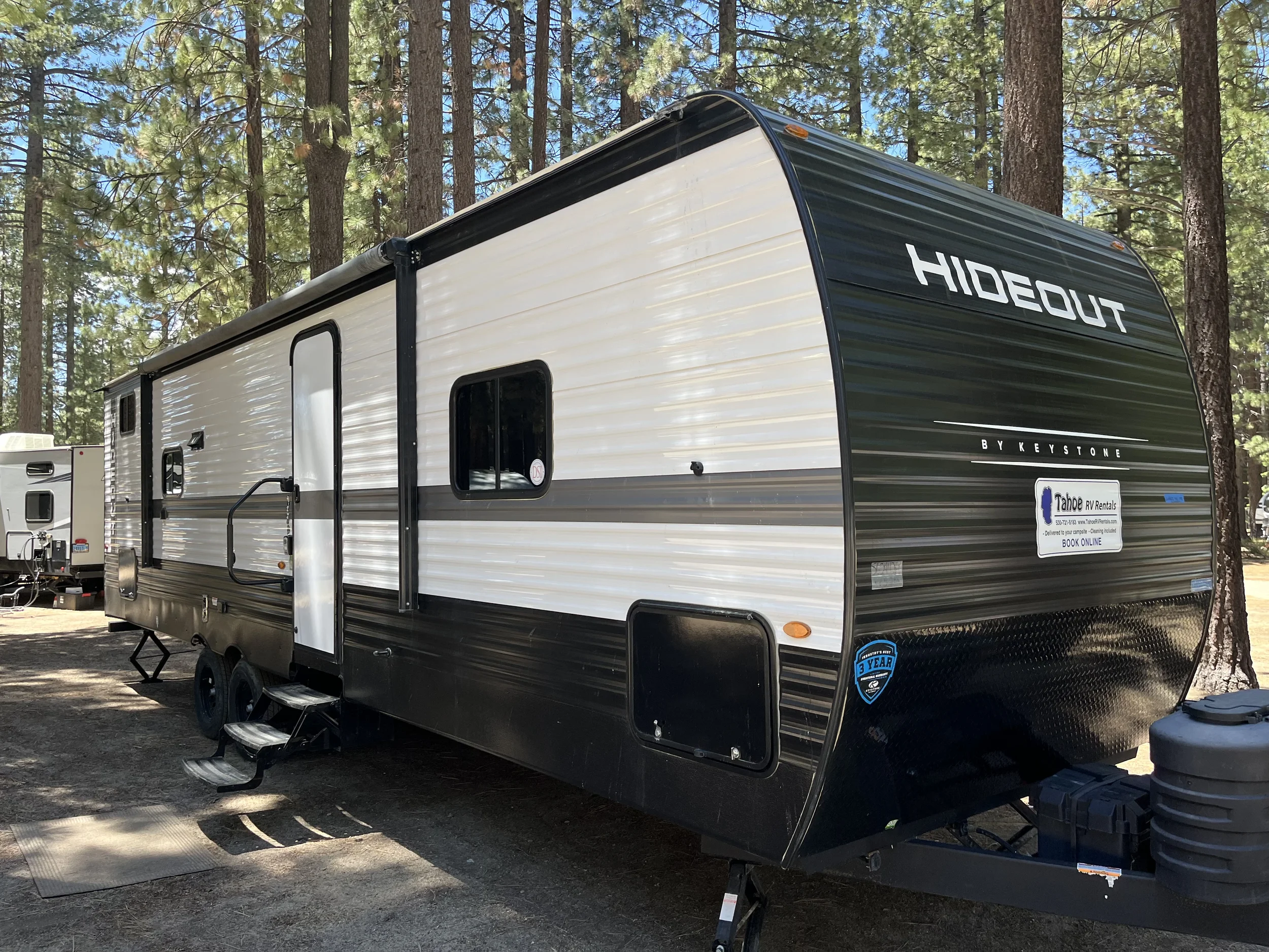 A modern black and white travel trailer named 'HIDEOUT' parked among tall pine trees at a campsite, with a set of portable steps leading to the door and a small sticker indicating a 3-year warranty on the bottom right corner.