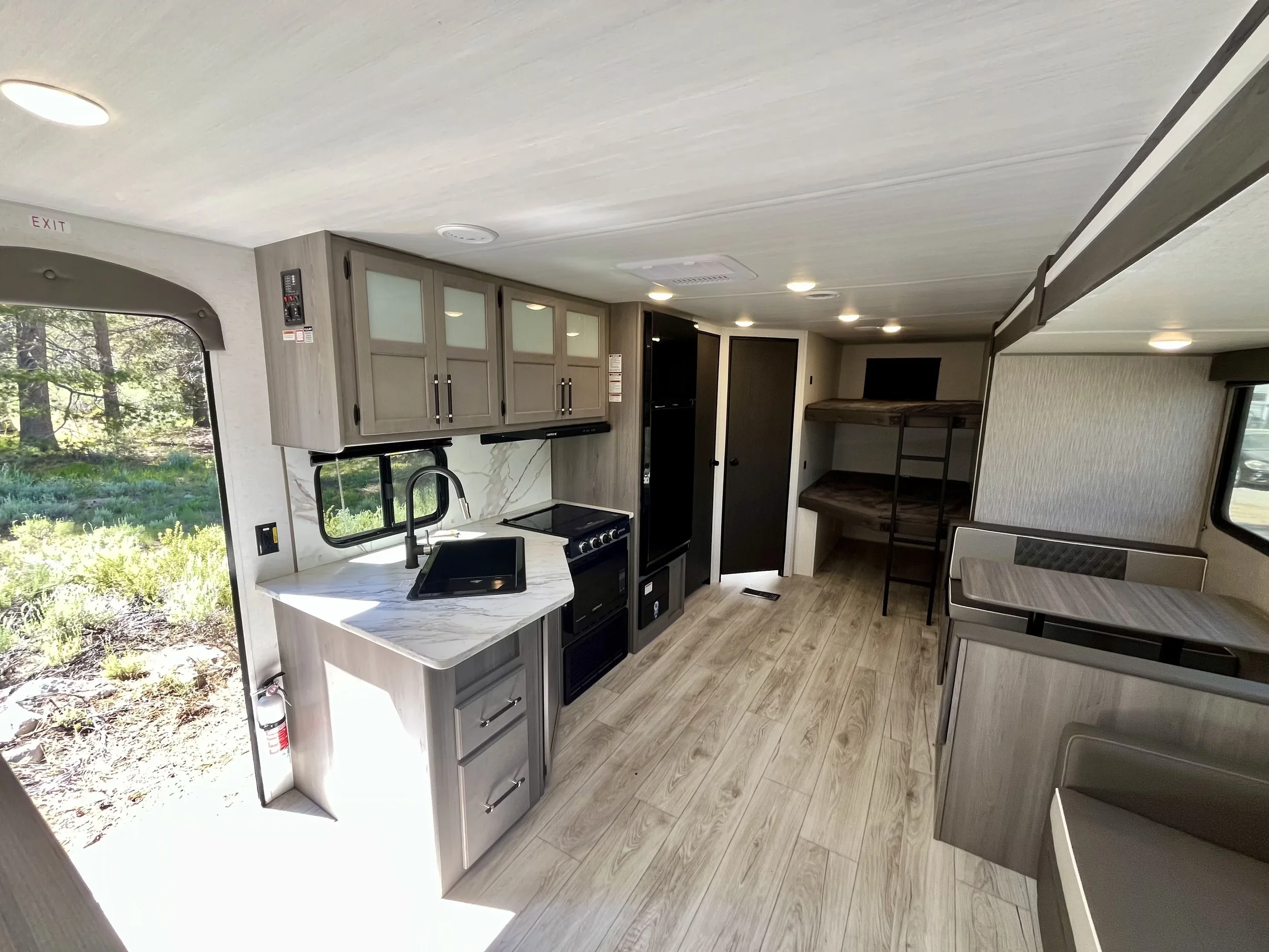 Interior of a modern RV with gray cabinets, a stove, sink, window, and dining area with a table and chairs.
