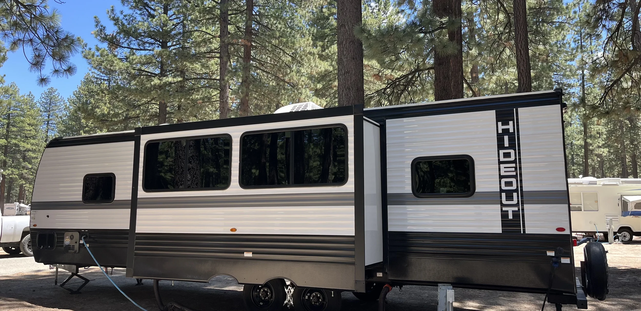 A white and black RV trailer parked among tall pine trees in a forest setting with a clear blue sky overhead.