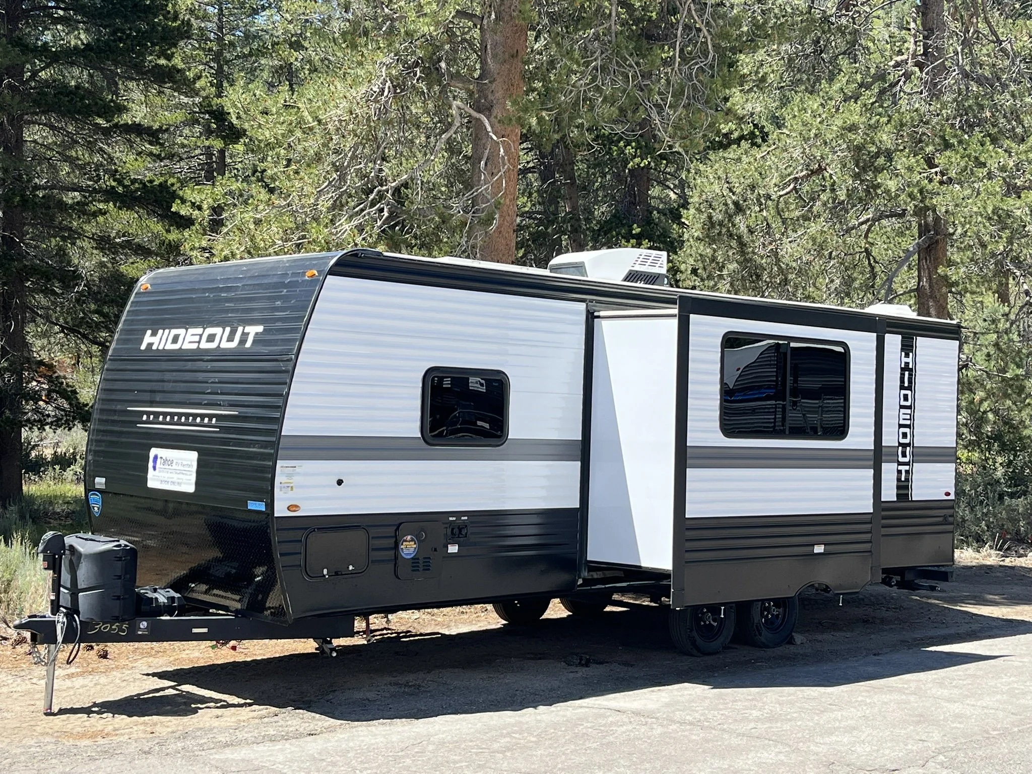 A modern travel trailer labeled 'HIDEOUT' parked on a dirt surface in a forested area with tall trees.