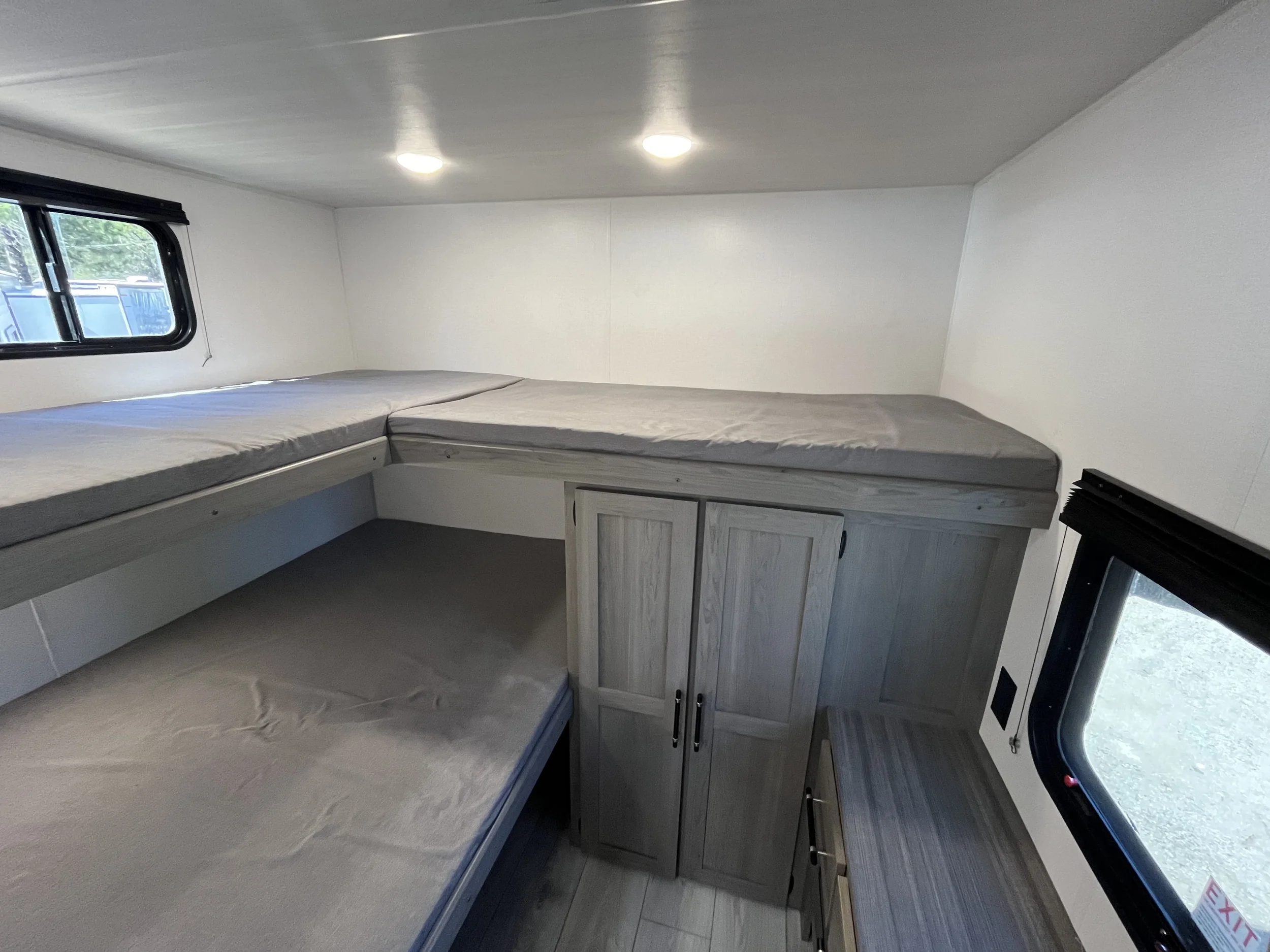 Interior of a small camper or RV with two bunk beds, a window on the side, a storage cabinet, and a small refrigerator, with ceiling lighting.
