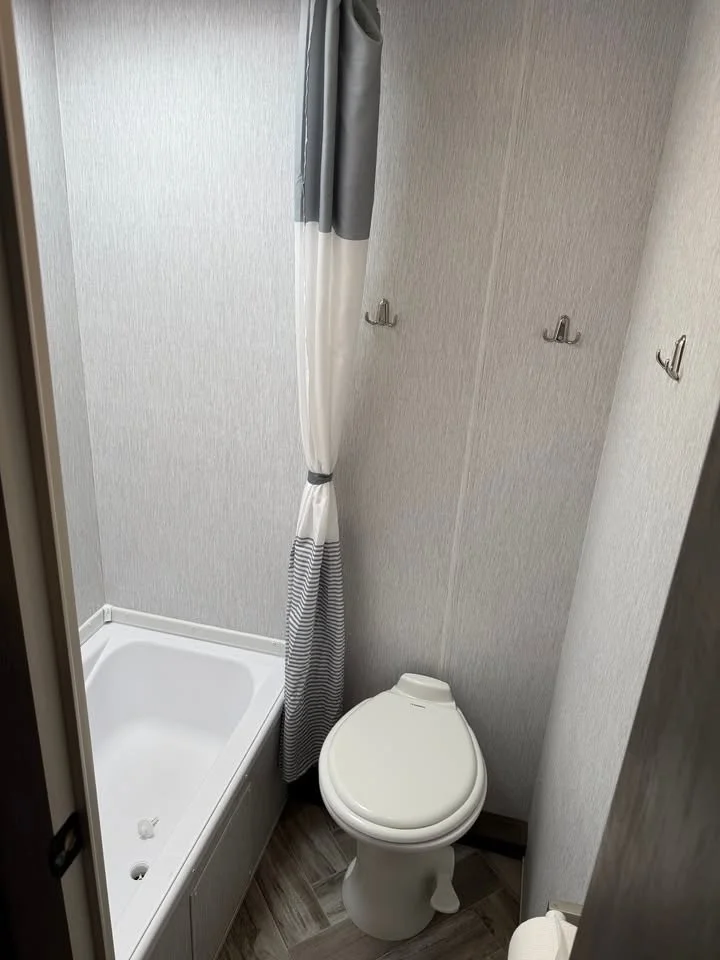 Small bathroom with a bathtub, a shower curtain, and a toilet. The walls are light-colored with a textured finish, and the floor has wood-like tiles.