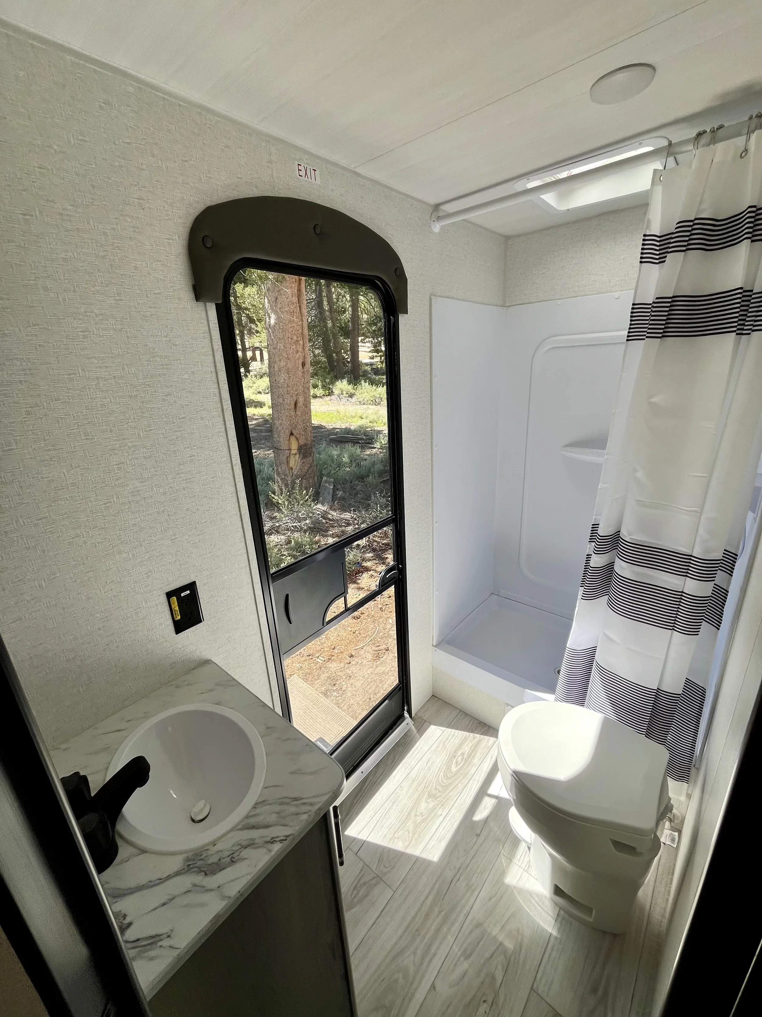 A small bathroom inside a camper or RV with a toilet, a sink with a marble countertop, a shower stall with a striped curtain, a window showing trees outside, and a door leading outside.