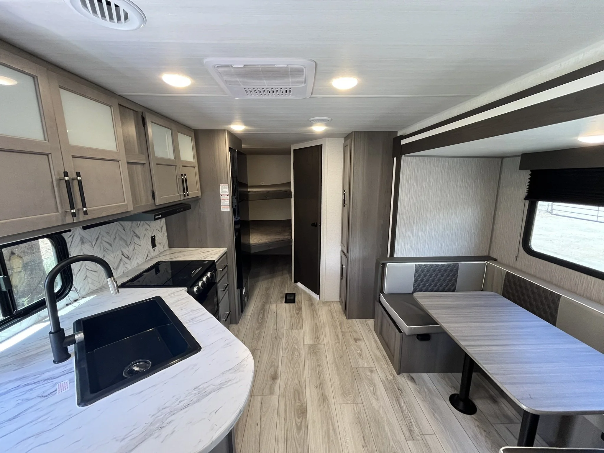 Interior view of a modern RV or camper with kitchen area, stove, sink, and a dining booth with a window, cabinetry, and ceiling lights.