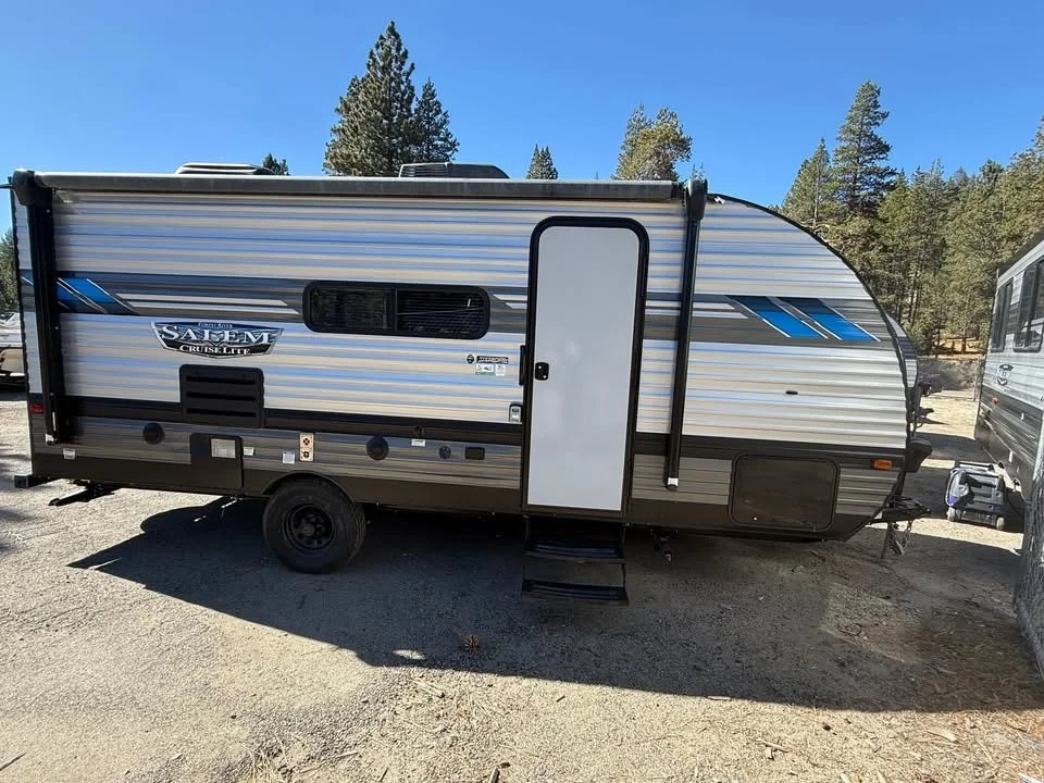 A grey and black travel trailer with blue accents parked on a dirt lot, surrounded by trees and a clear blue sky.