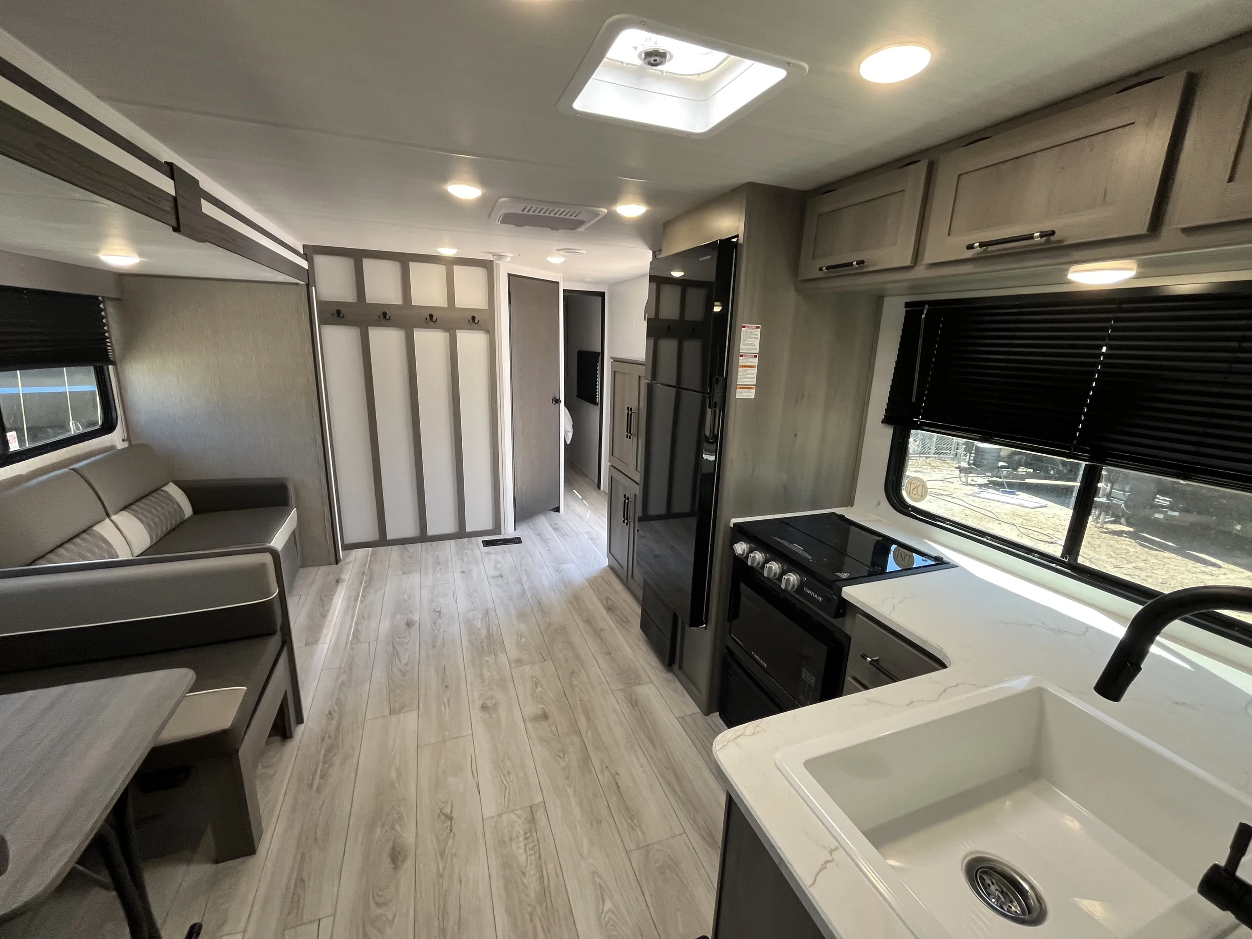 Interior of a modern travel trailer or RV with a small kitchen area, black cabinets, a stove, and windows with blinds, beige sofa, and light wood flooring.