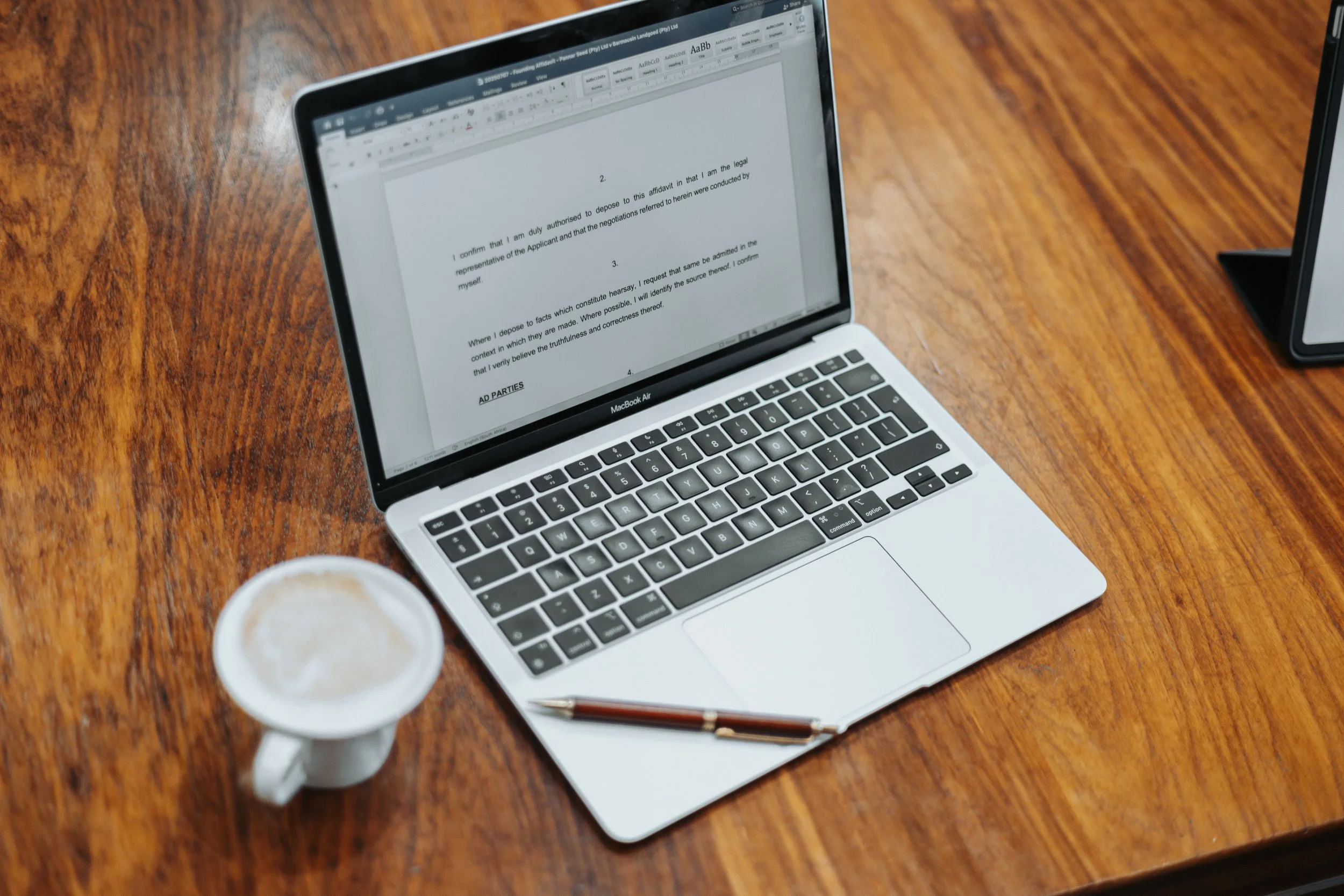 A silver MacBook Air laptop on a wooden table with a document open on the screen, next to a cup of coffee, a pen, and a tablet.