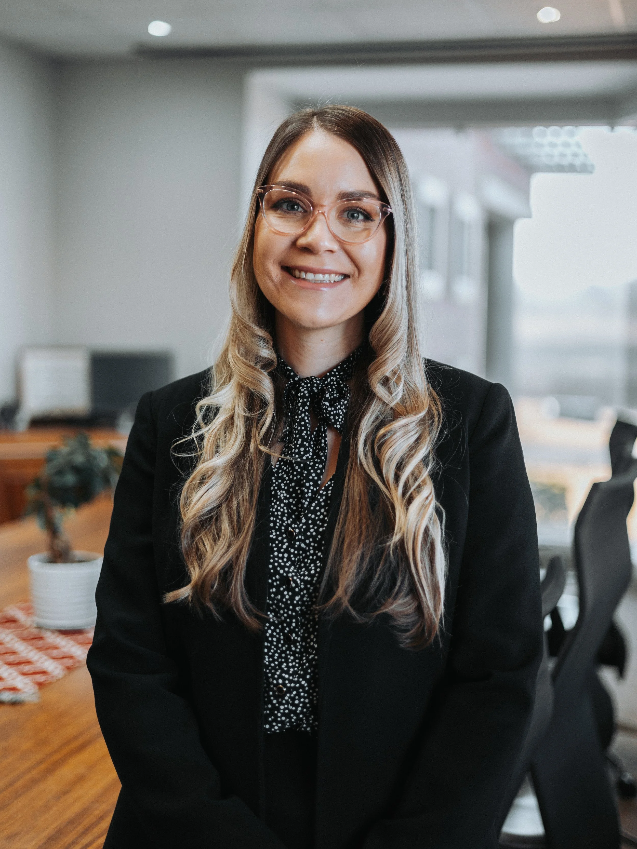 A smiling woman with long, wavy hair, wearing glasses, a black blazer, and a polka-dot blouse, in an office setting.