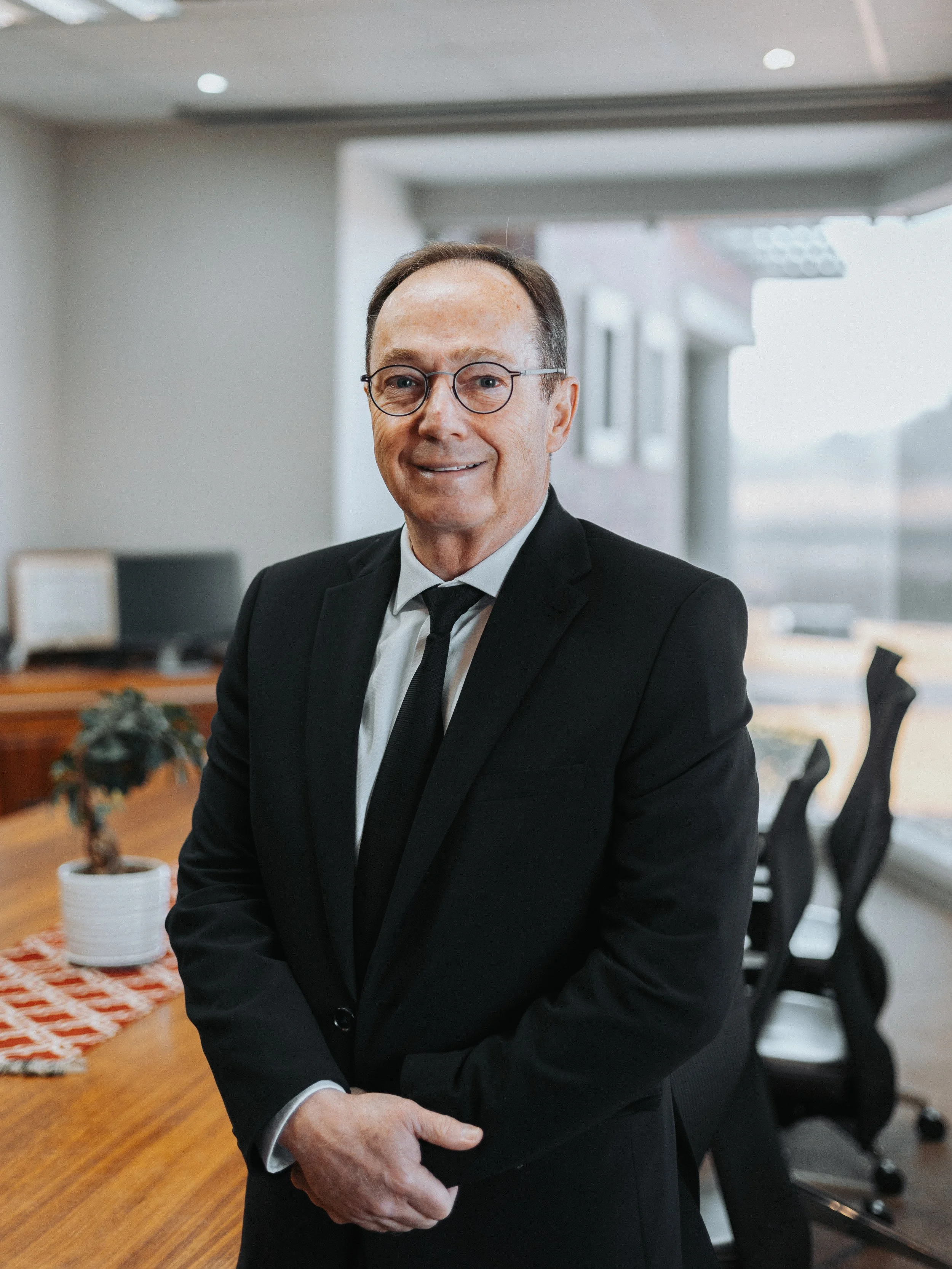 A man dressed in a black suit with a white shirt and black tie, wearing glasses, sitting in an office with wooden furniture and a potted plant in the background.