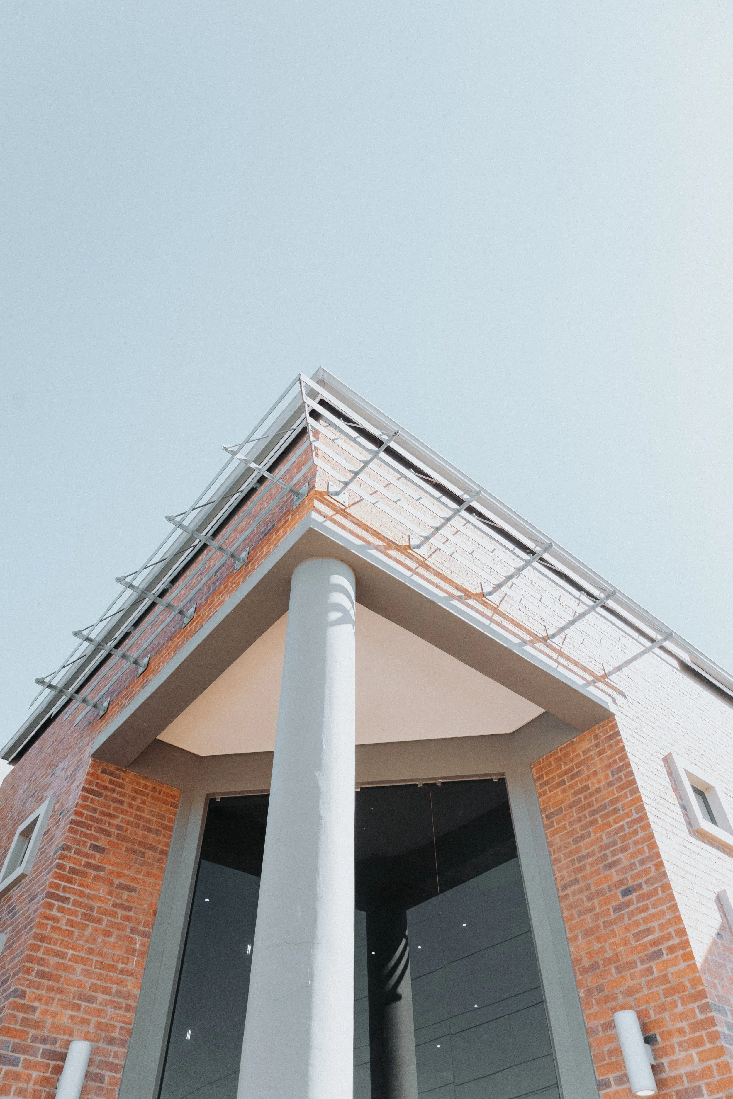 Close-up of a modern building corner with brick, gray metal accents, a glass entrance, and a large white column support, against a clear sky.