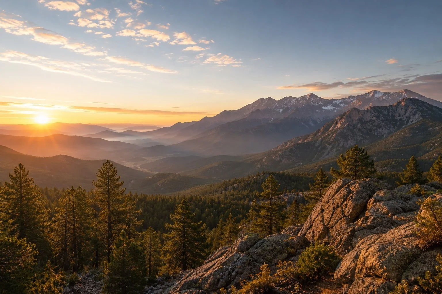 Sunset over a mountain range with pine trees and rocky foreground.