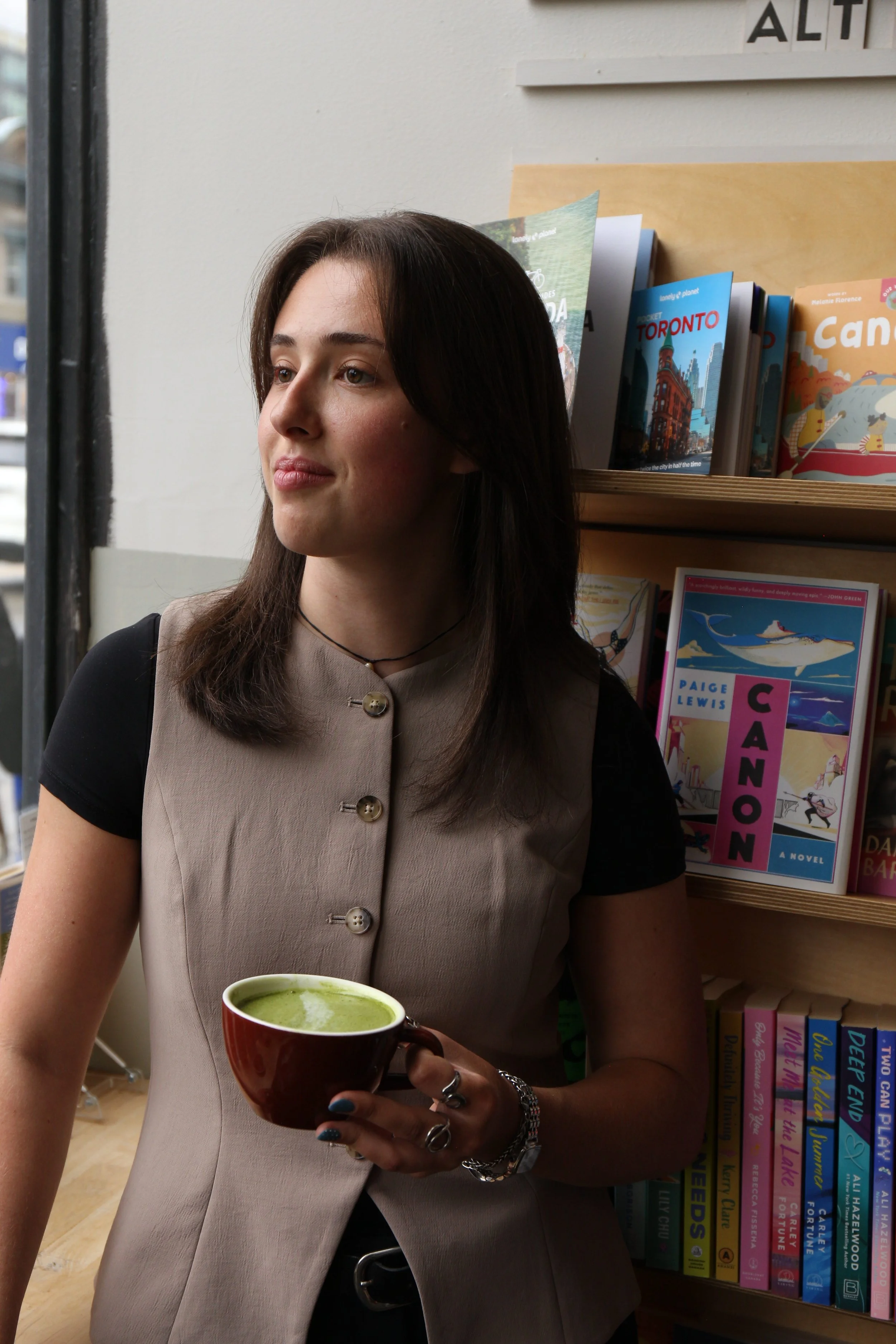 A young woman with dark brown hair, wearing a beige vest with buttons and a black shirt with short sleeves, is holding a brown mug filled with green tea or matcha. She has rings on her fingers and multiple bracelets on her wrist. Behind her, there is a bookshelf filled with colorful books about travel destinations and children's books, and a window on the left side of the image.