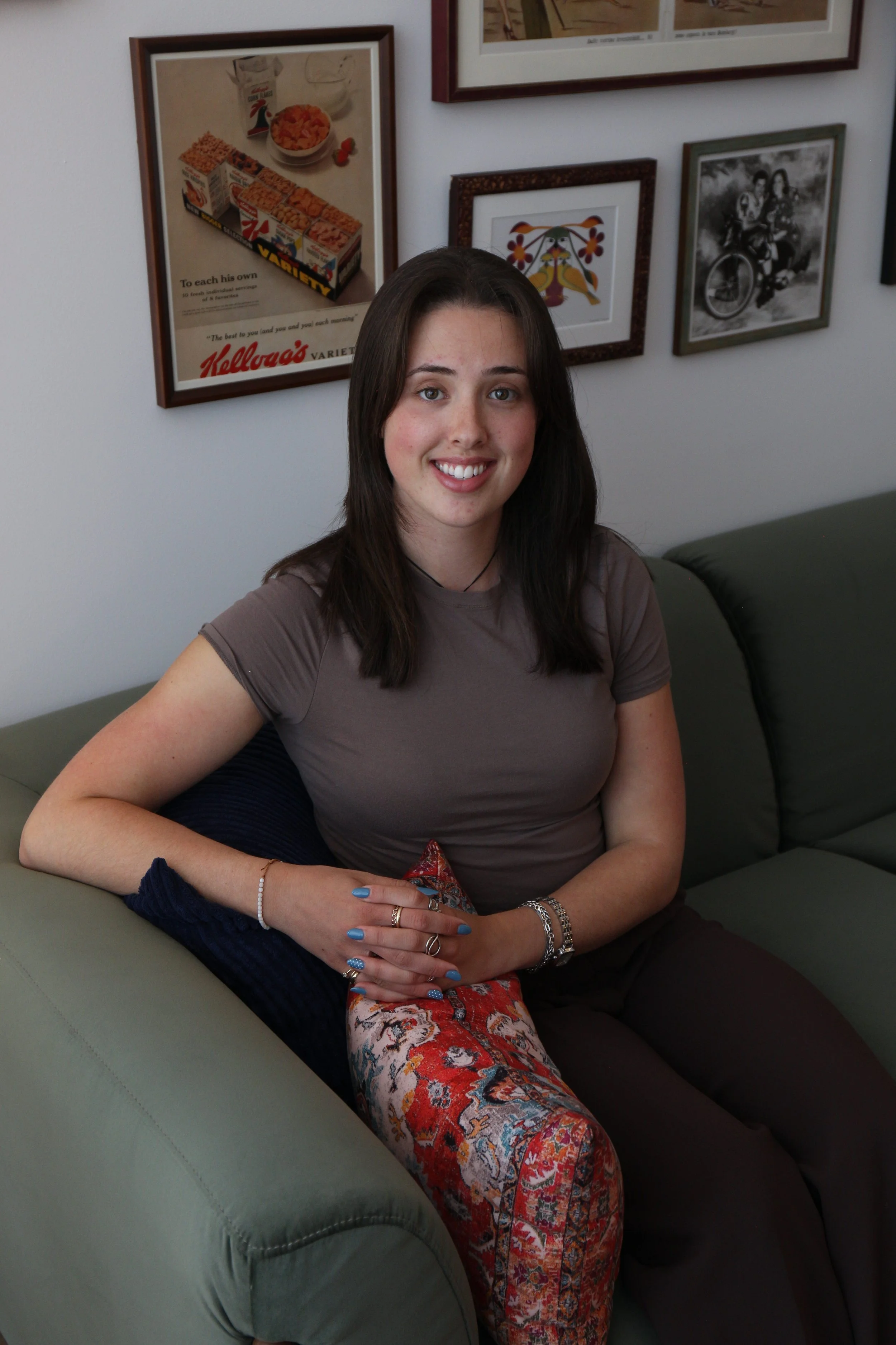 A young woman with long dark hair smiling and sitting on a green sofa with a tapestry and various framed pictures on the wall behind her.