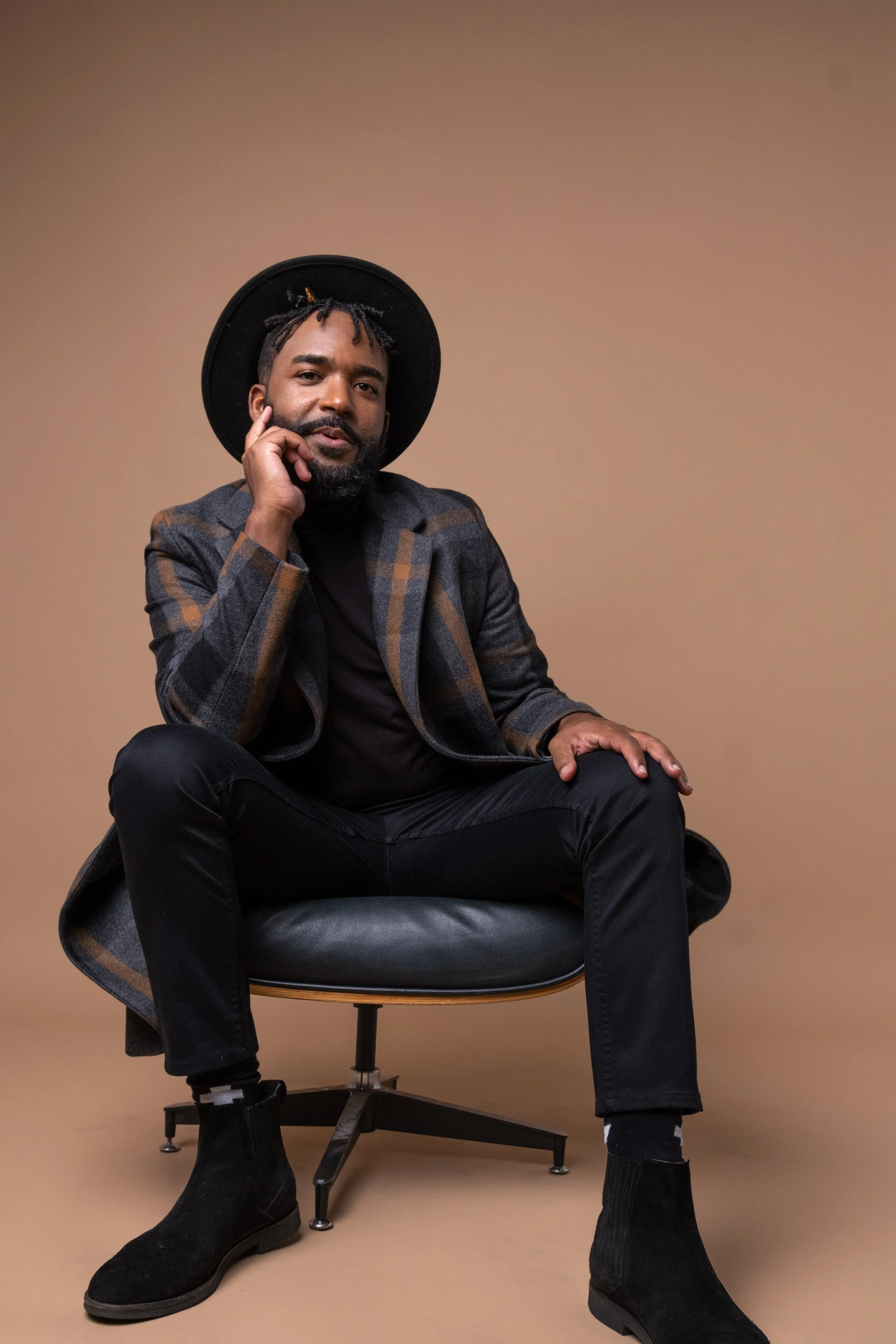 A man with a beard and dreadlocks wearing a black hat, plaid blazer, black turtleneck, black pants, and black boots, sitting in a black leather chair against a beige background.