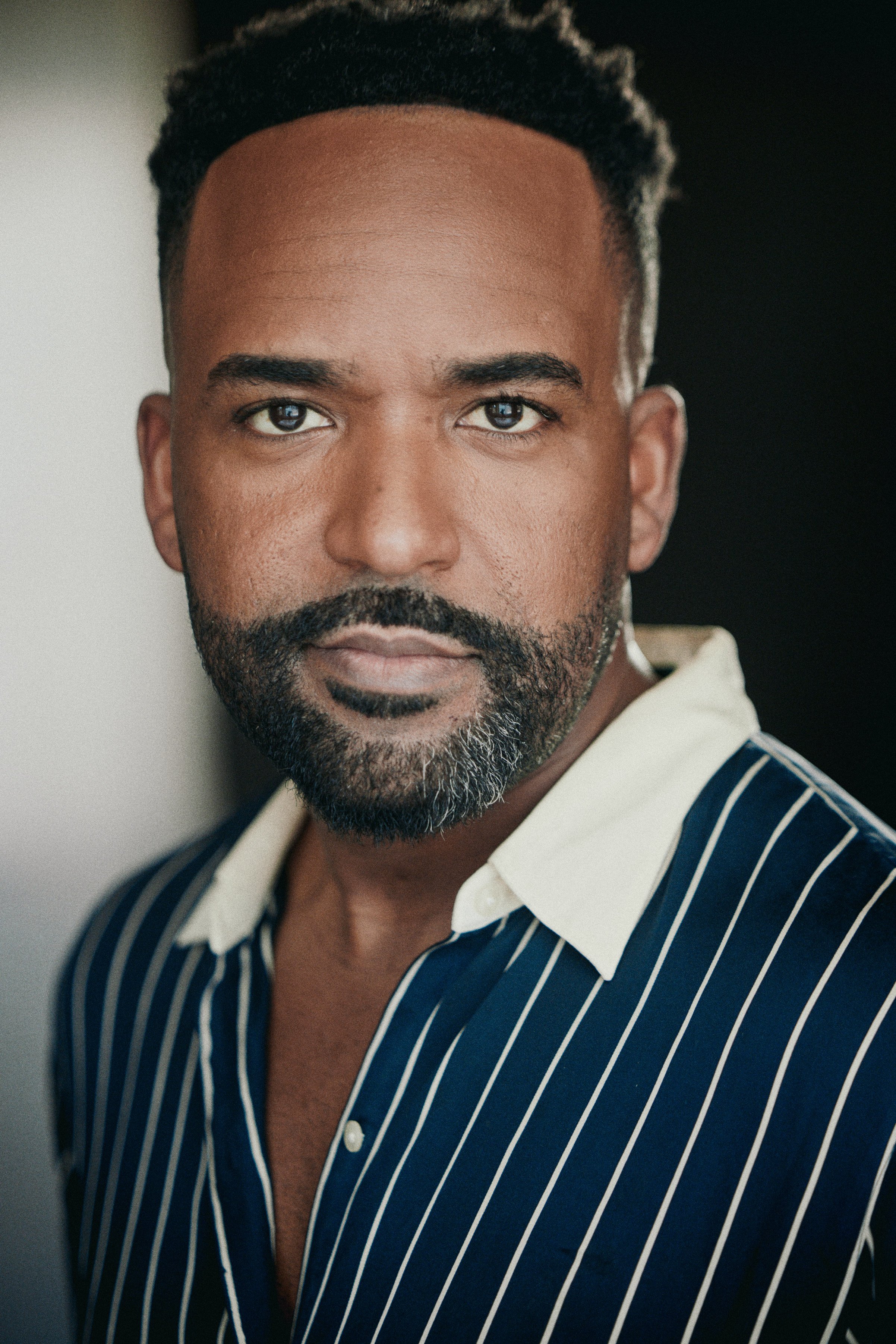 A close-up portrait of a man with dark skin, short black hair, a beard, and wearing a blue and white striped shirt.