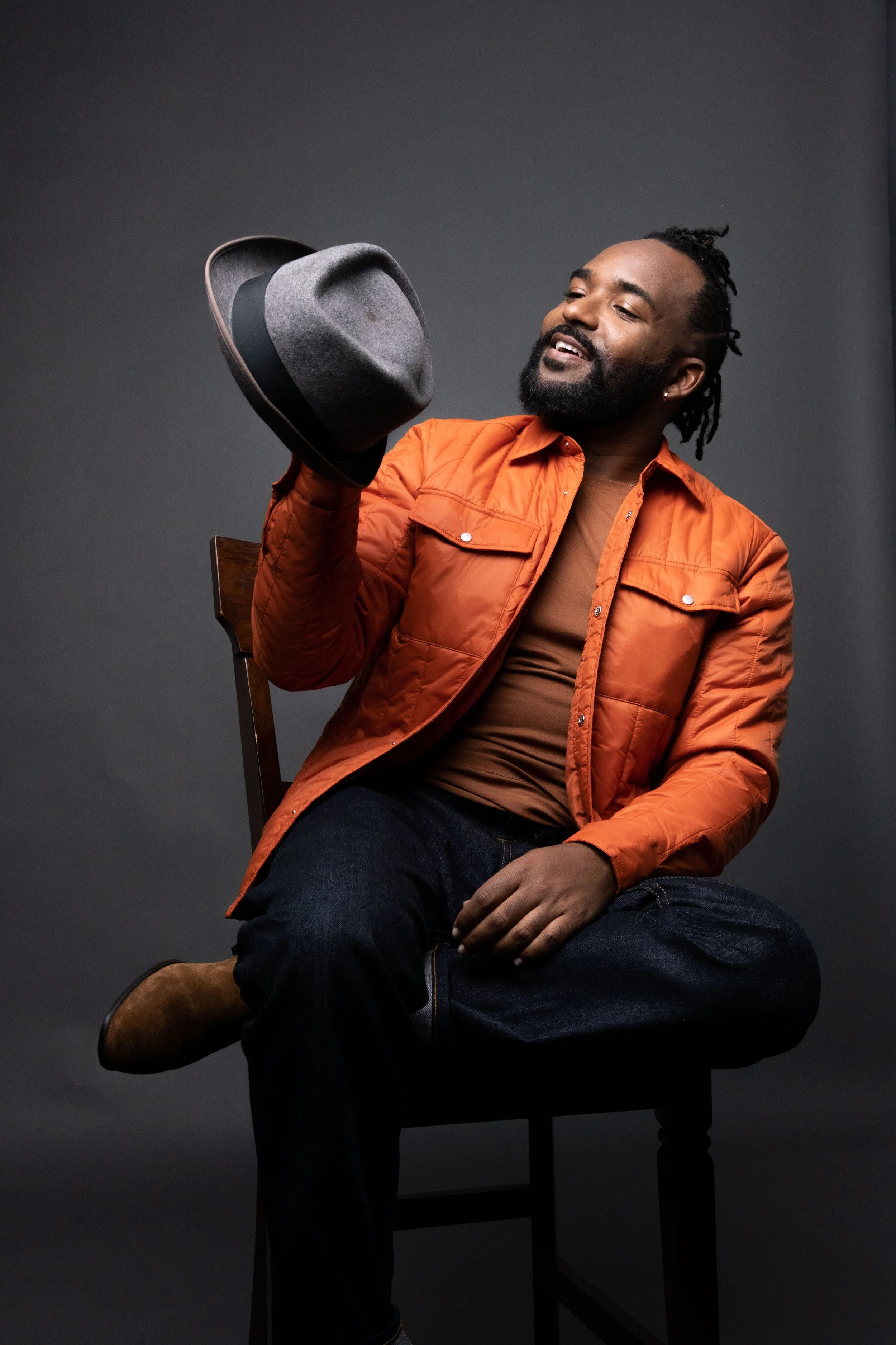 A man with a beard and dreadlocks wearing an orange jacket and brown shirt, sitting on a dark wooden chair, holding a gray fedora hat in his right hand, against a gray background.
