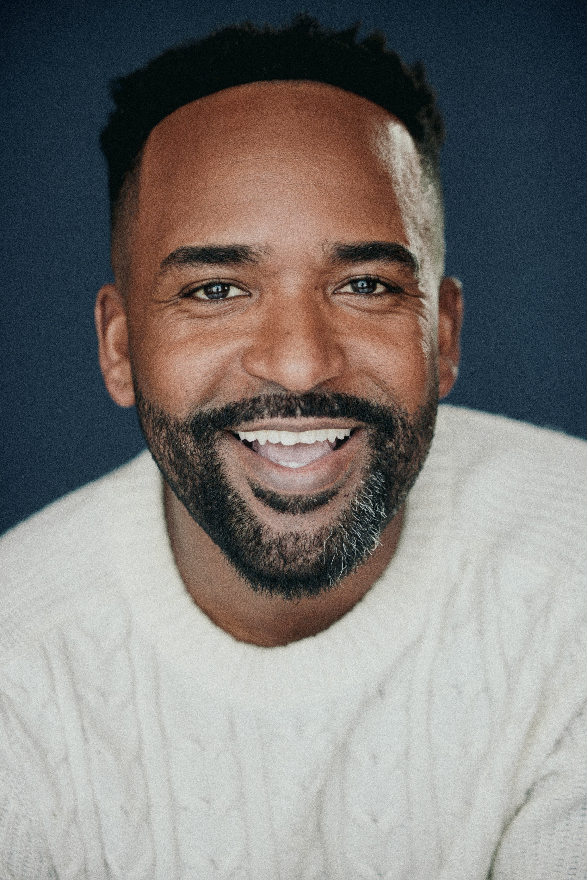 Close-up portrait of a smiling Black man with a beard, wearing a light-colored cable-knit sweater, against a dark blue background.
