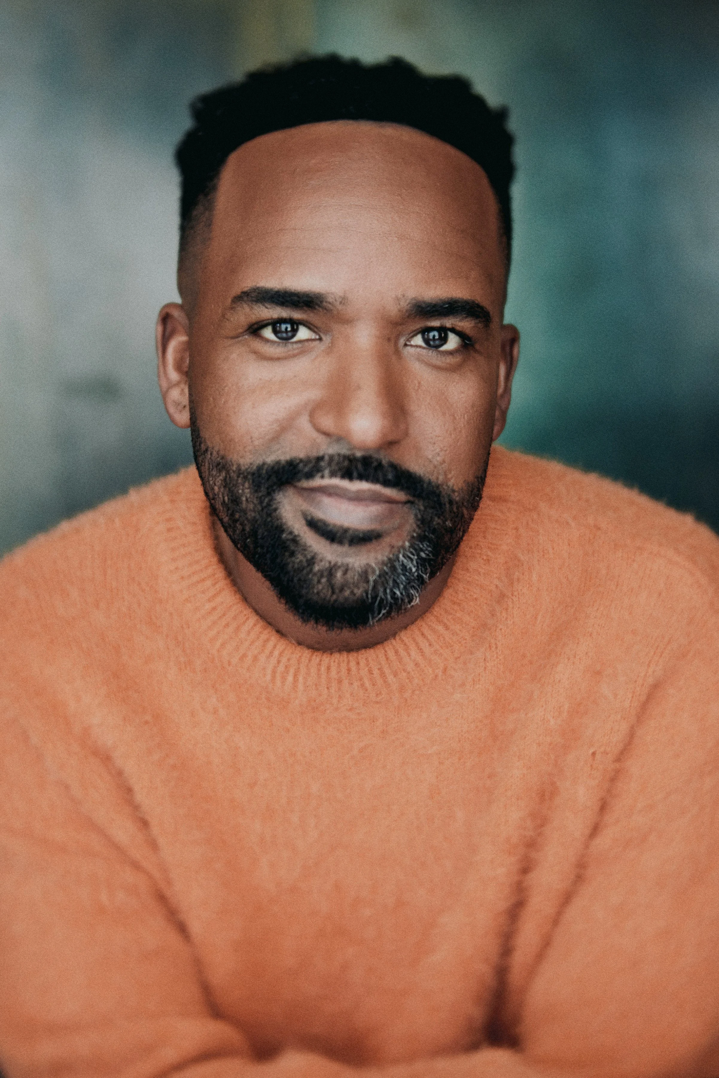 A close-up portrait of a man with dark skin, short black hair, a beard, and a mustache, wearing an orange sweater, with a neutral background.