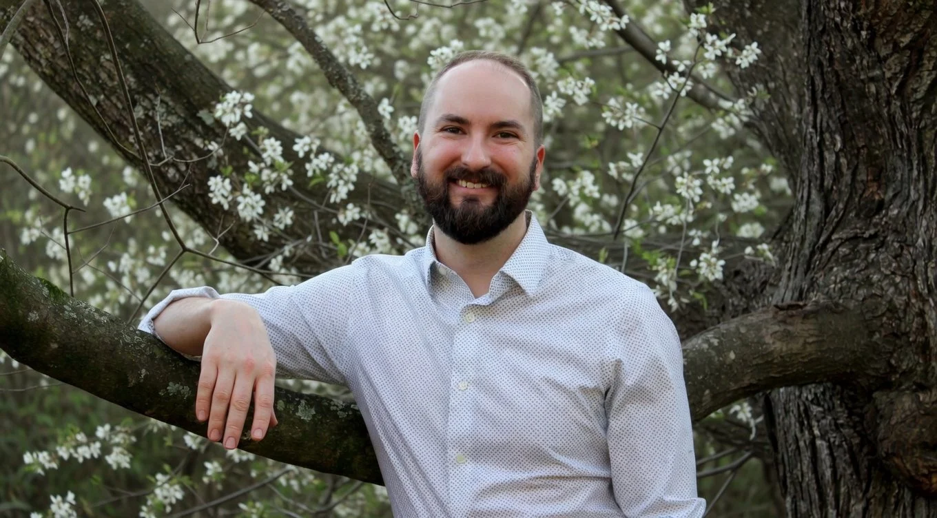 A smiling man with a beard and short hair, wearing a light-colored, button-up shirt, leaning on a tree branch with blooming white flowers in the background.