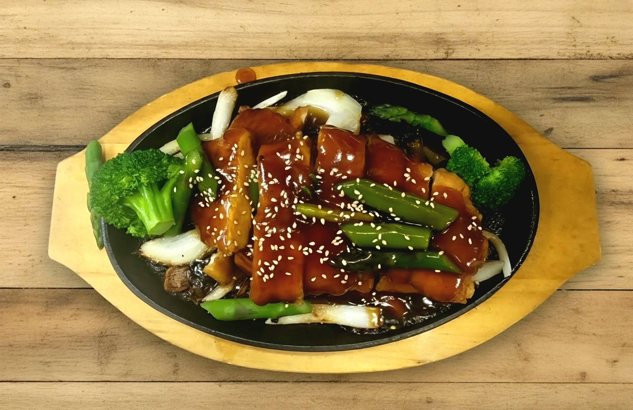 A sizzling hot plate of stir-fried vegetables and grilled chicken topped with teriyaki sauce, sprinkled with sesame seeds, and garnished with broccoli, green beans, and onions, served on a wooden tray.