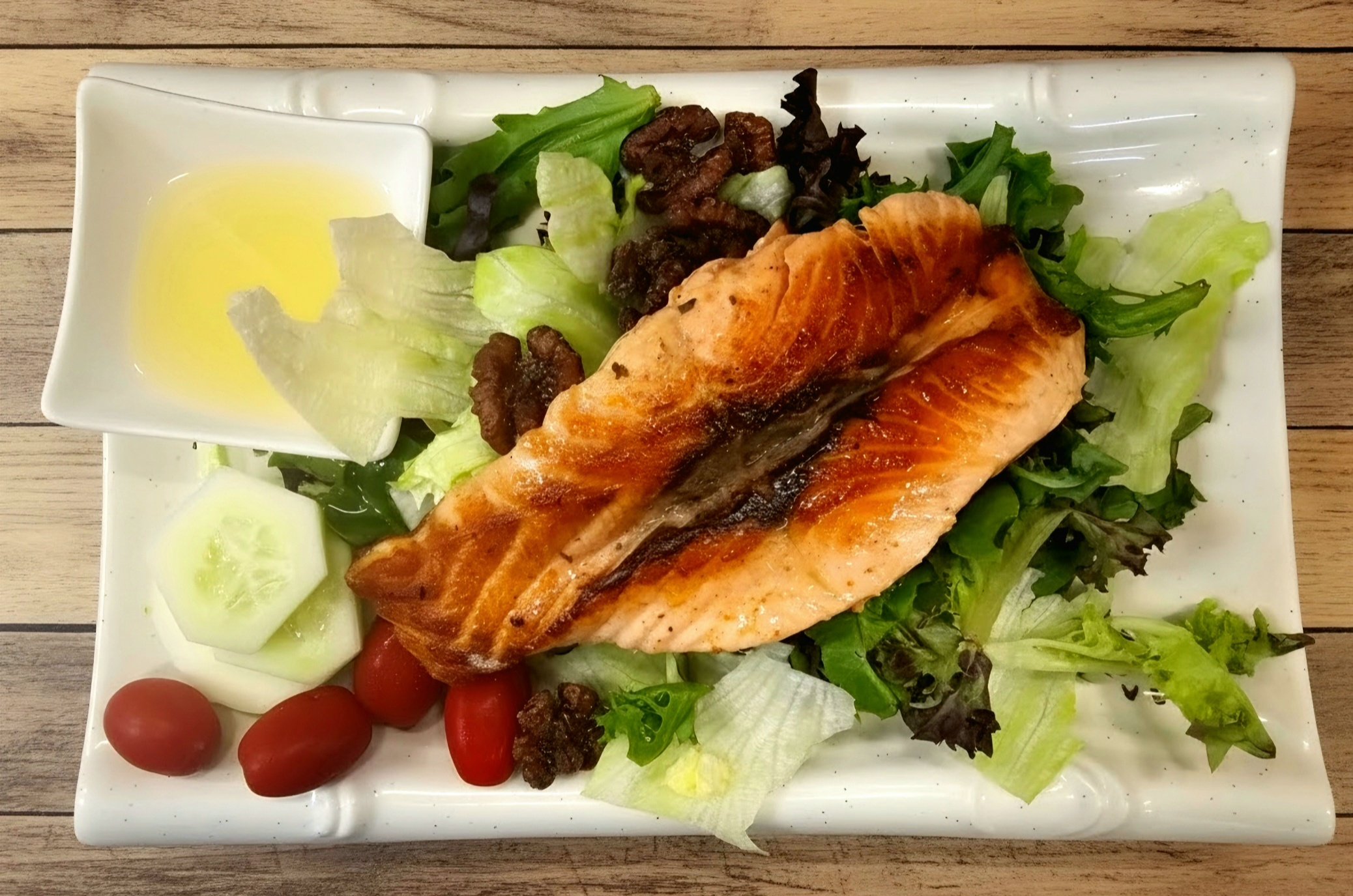 Plate with grilled salmon, mixed greens salad with cherry tomatoes, cucumber slices, bacon bits, and a small dish of dressing on the side.