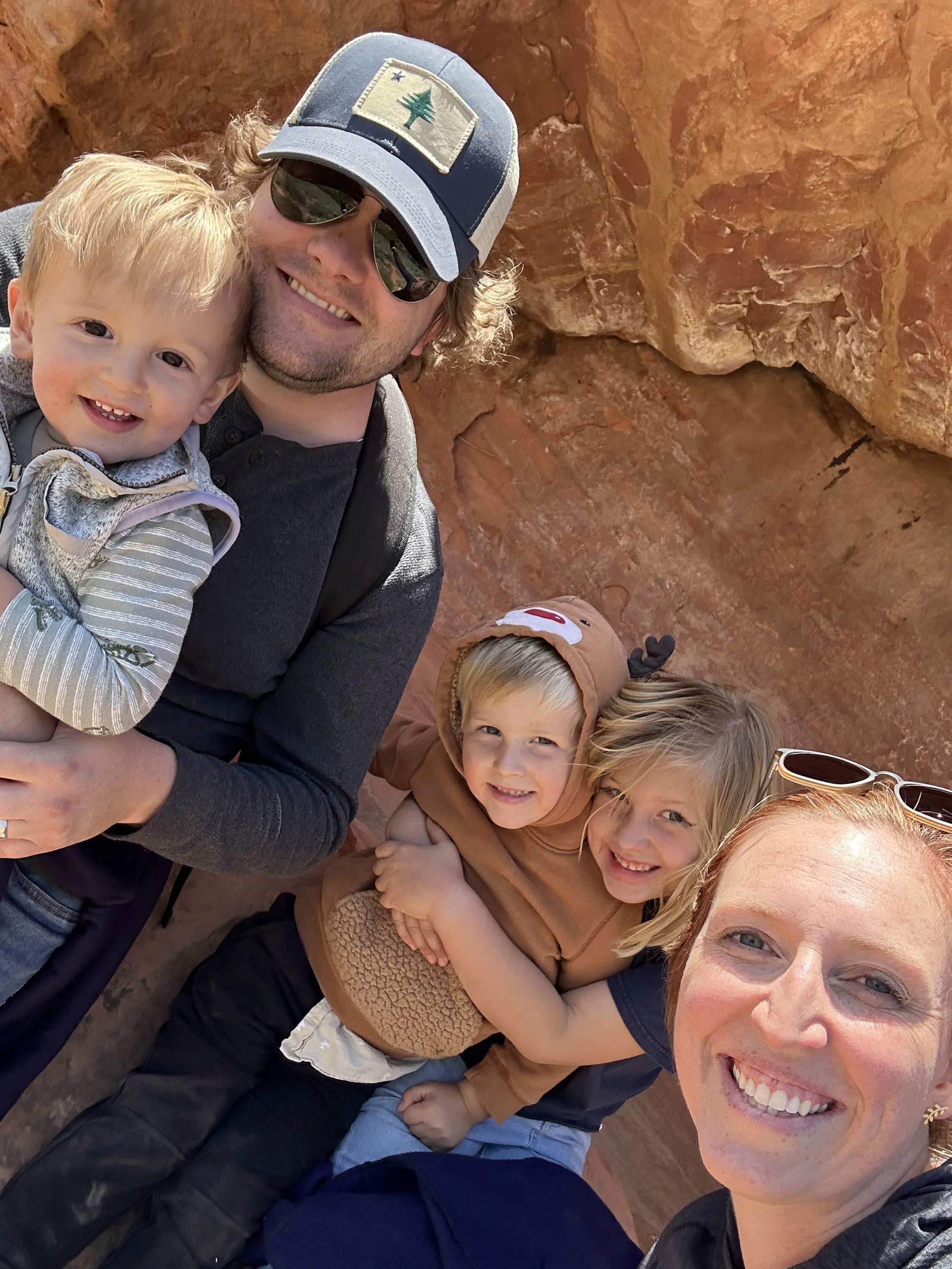 A group of smiling people, including a woman, a man wearing sunglasses and a cap with a tree emblem, and three children, taking a selfie together against a reddish rock formation.