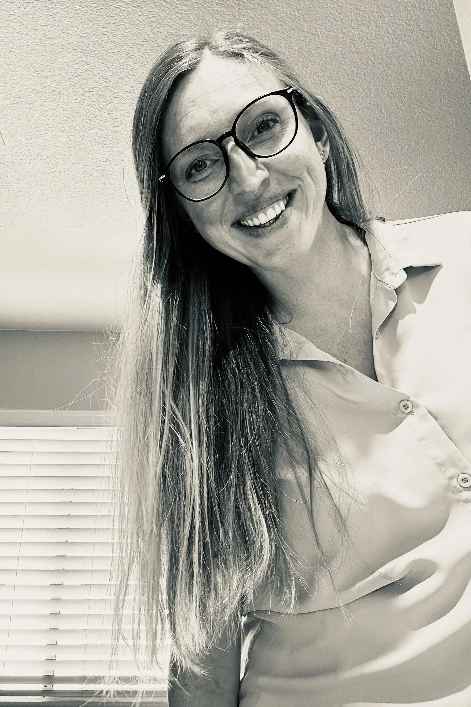 Black and white photo of a woman with long hair, wearing glasses and a collared shirt, smiling at the camera.