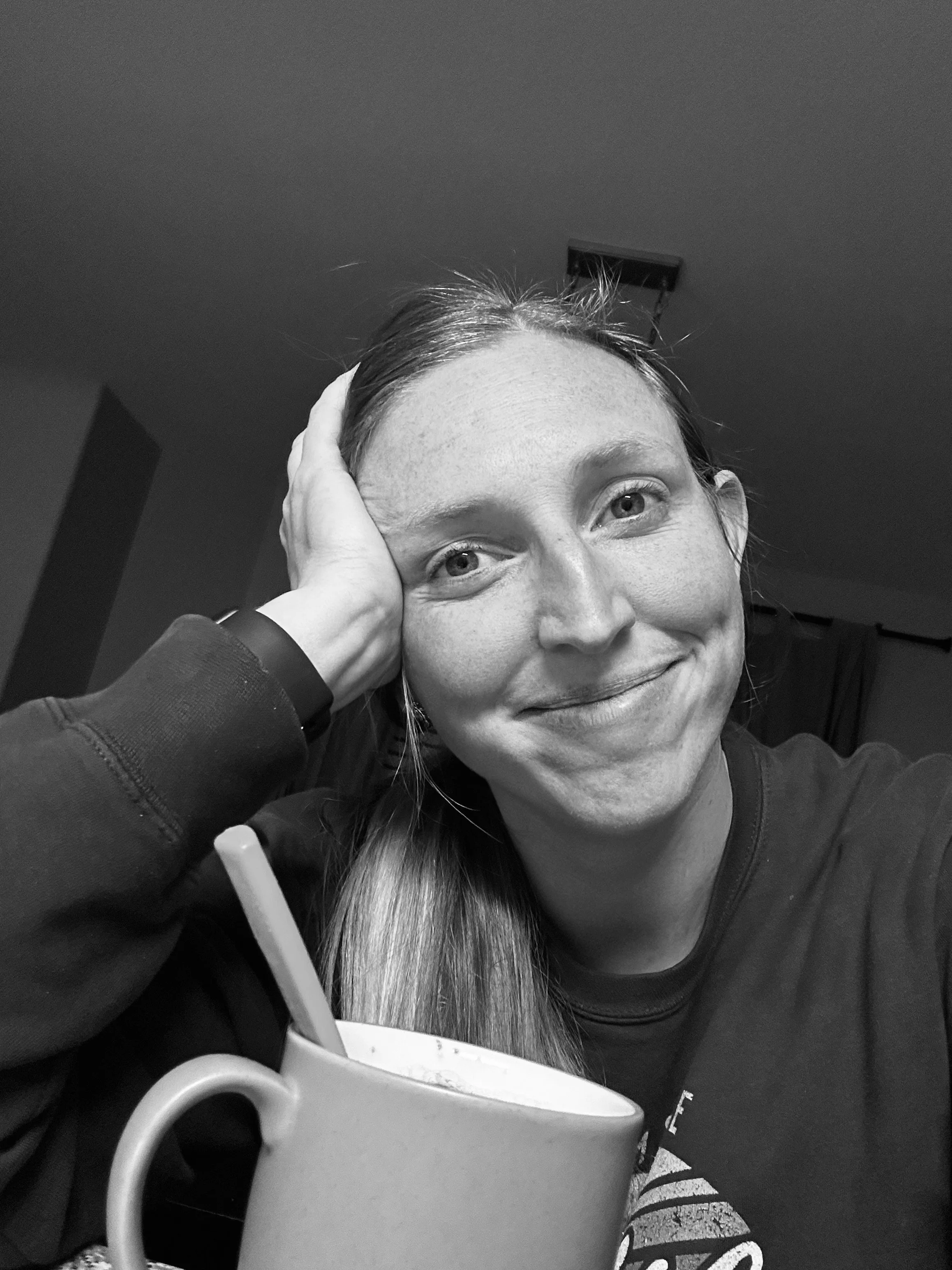 Black and white photo of a smiling woman with her hand resting on her forehead, sitting at a table with a mug and a pen in the foreground.