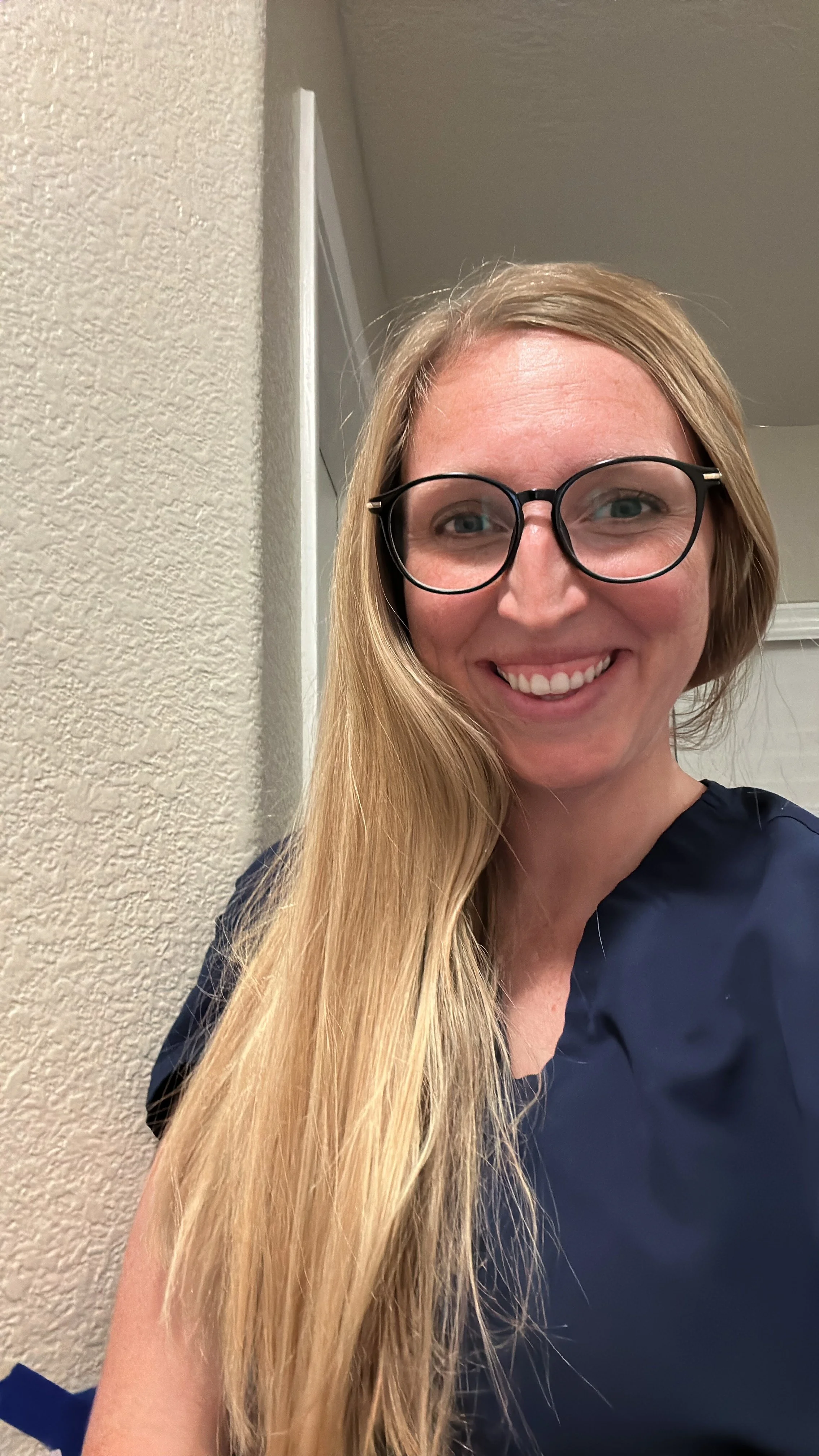 A woman with long blonde hair, wearing glasses and a dark blue shirt, smiling at the camera.