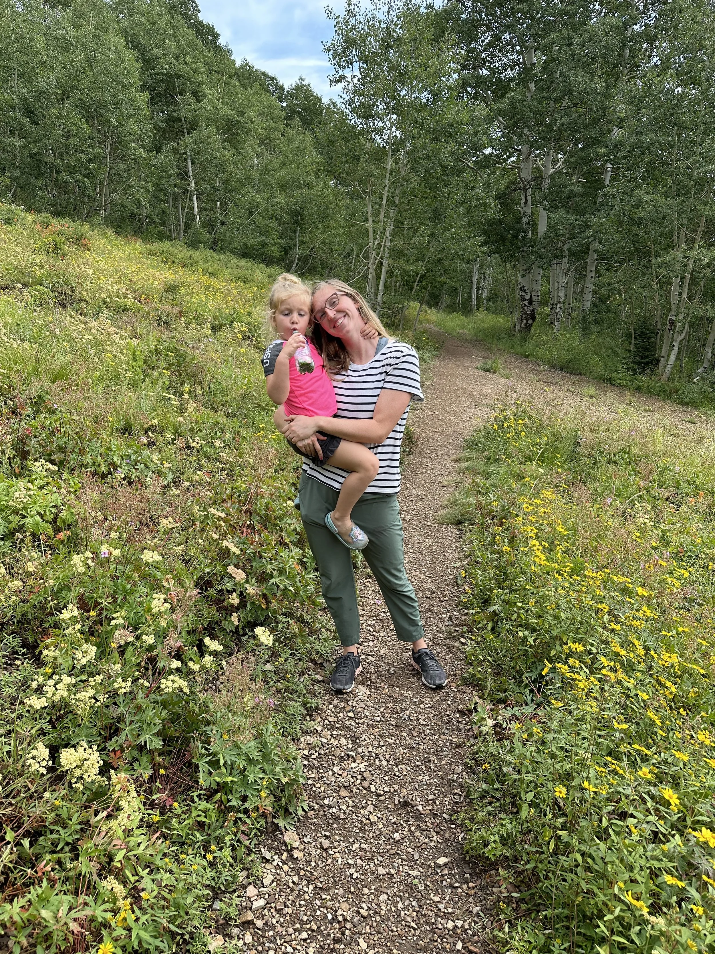 A woman with glasses and a striped shirt is holding a young girl with blonde hair and a pink shirt, both smiling, on a nature trail surrounded by green trees and wildflowers.