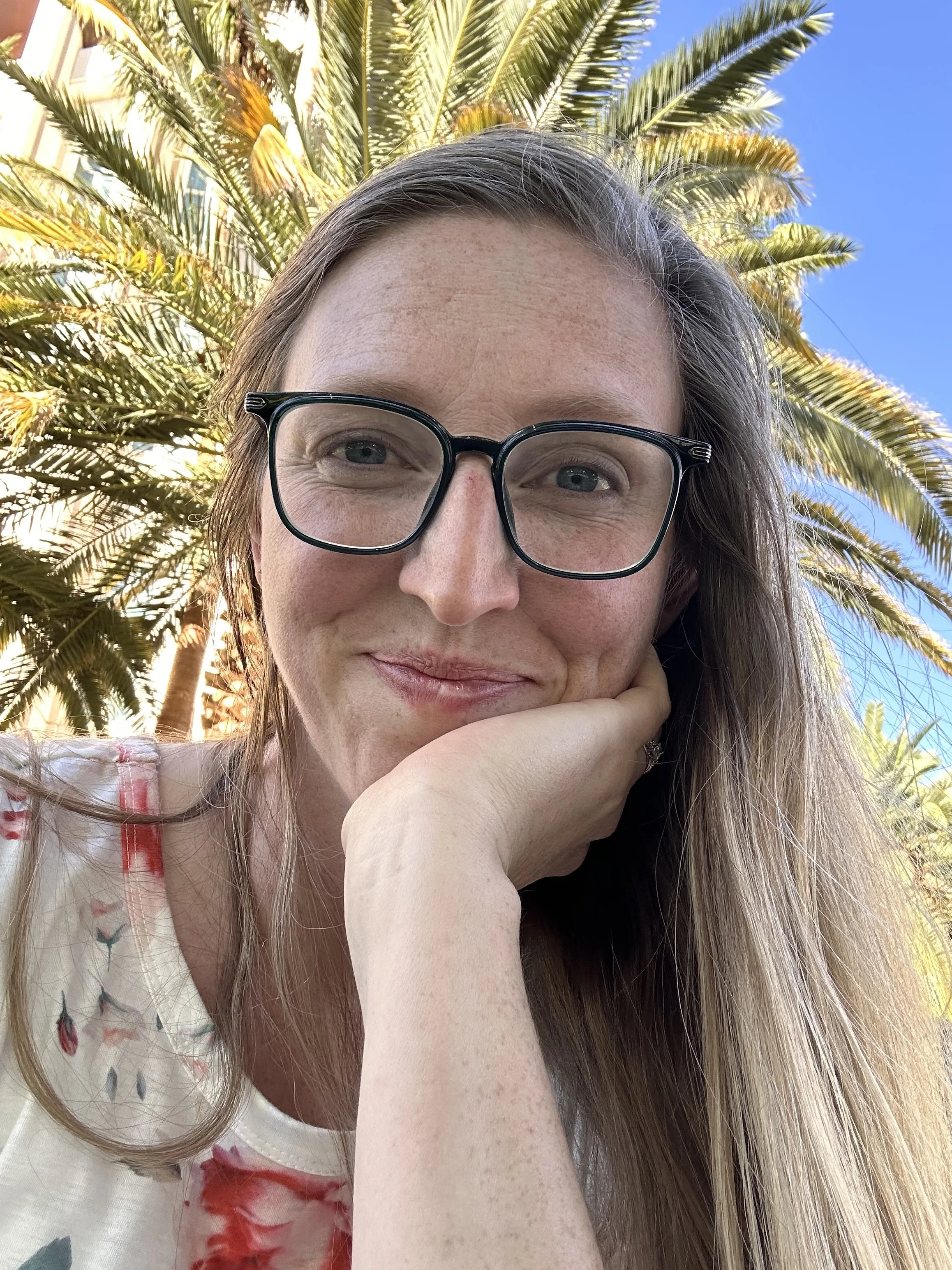 A woman with glasses and long hair taking a selfie outdoors. She is resting her chin on her hand, with iguana-patterned clothing, in front of a palm tree and bright blue sky.