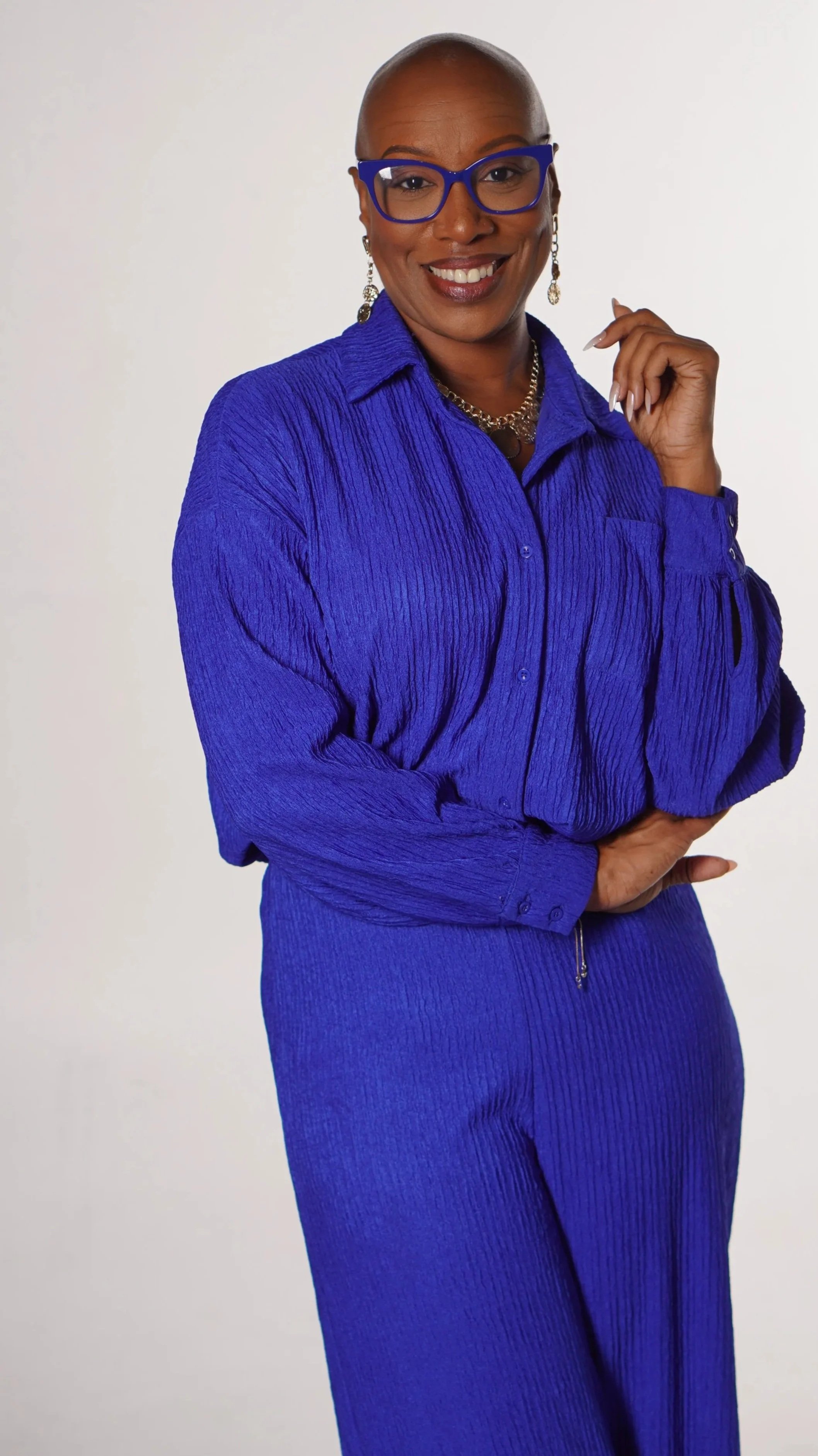 A woman with a bald head, wearing blue glasses, a blue blouse, and matching blue pants, smiling and standing with her hand near her face against a white background.