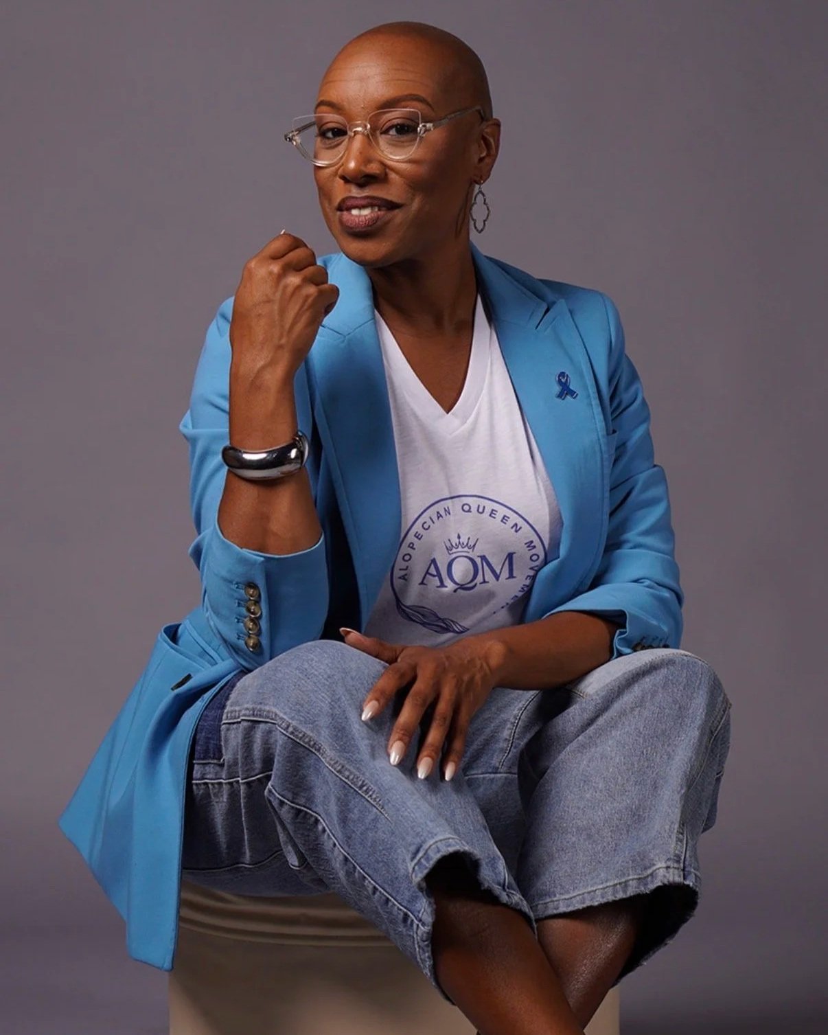 A woman with glasses, wearing a blue blazer and white T-shirt with a logo, seated with one leg crossed over the other, smiling at the camera against a gray background.