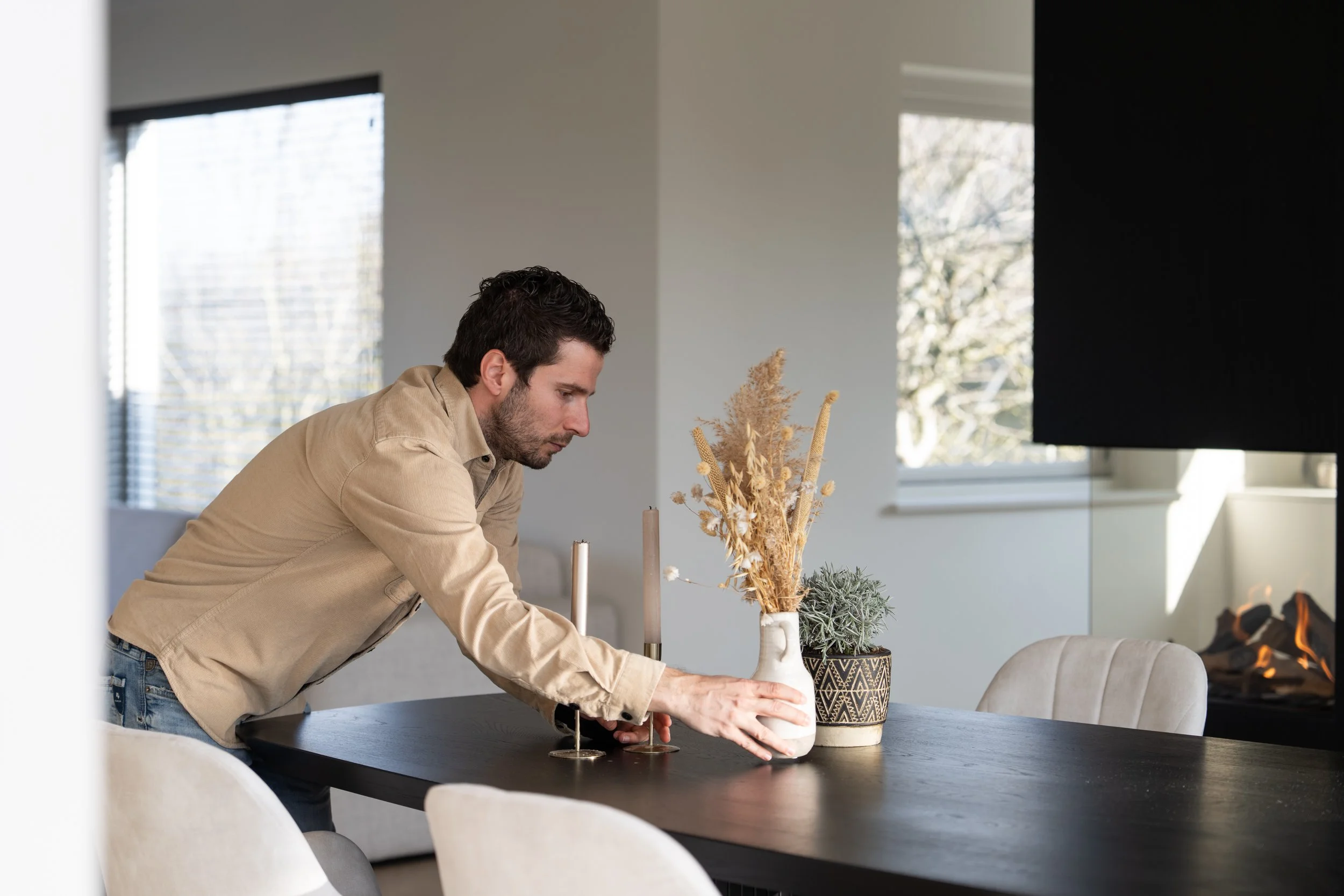 A man arranging decorative vases with dried flowers on a dining table near a window.