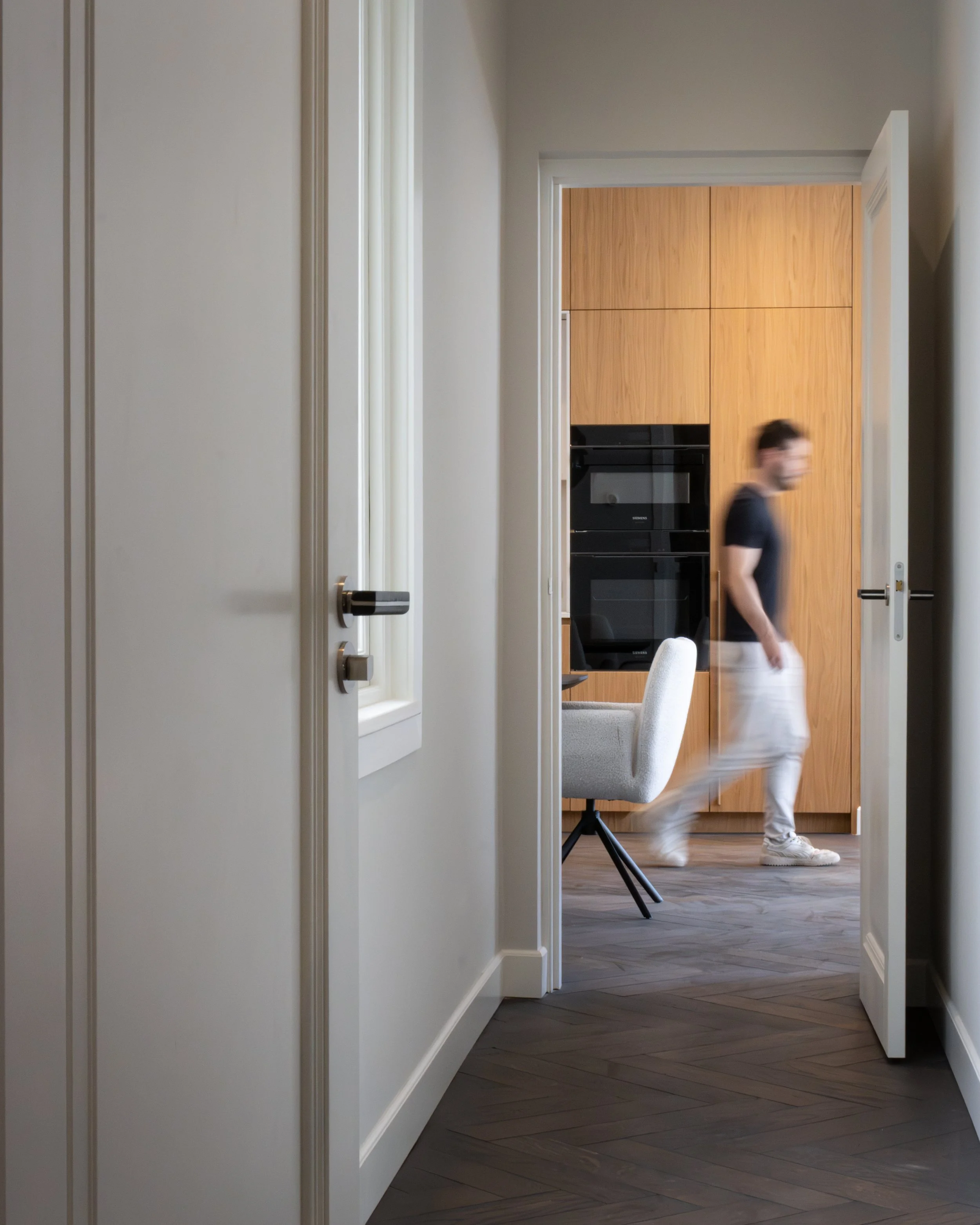 A corridor leading into a room with gray walls, a left door closed, and a wooden cabinet with black appliances in the room. A blurred person wearing a black t-shirt and light pants walks through the room.