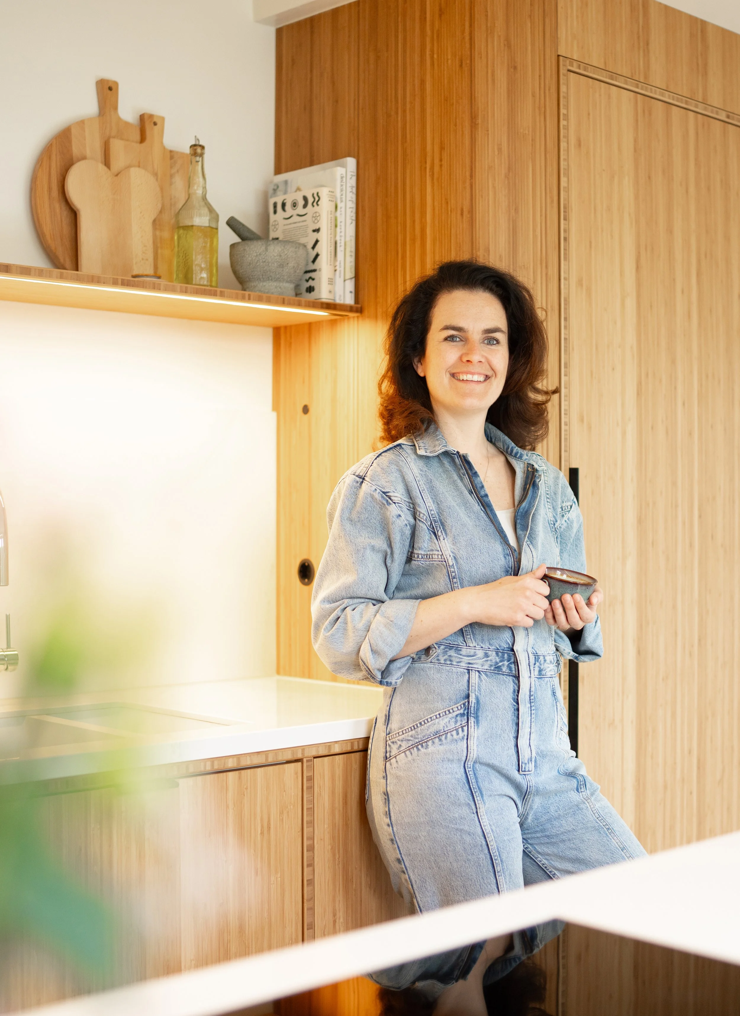 A woman with dark, curly hair smiles while holding a small bowl in a modern kitchen with wood cabinetry and a white countertop.