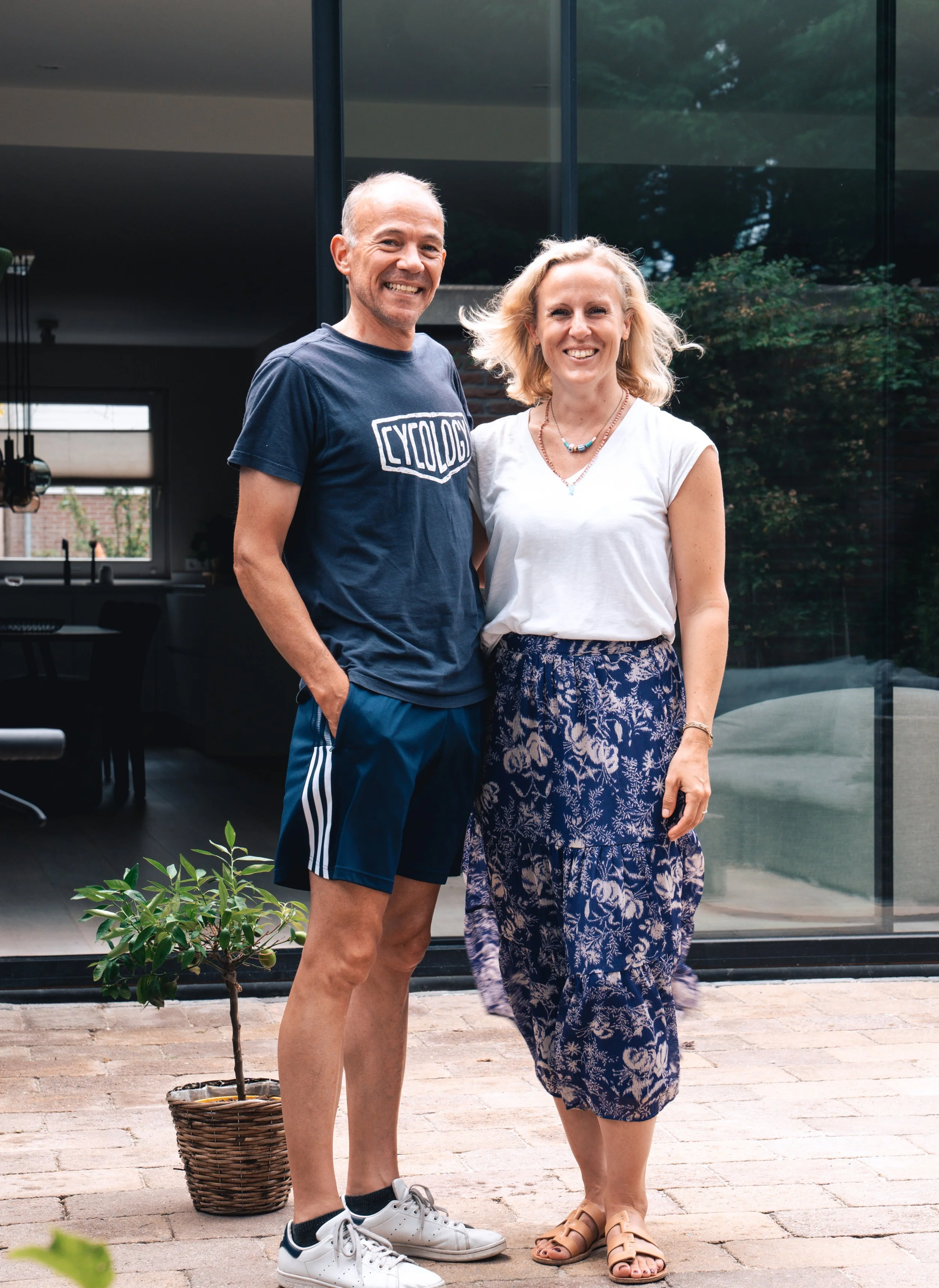 A man and woman standing outdoors in front of a glass building, smiling at the camera. The man is wearing a navy T-shirt with 