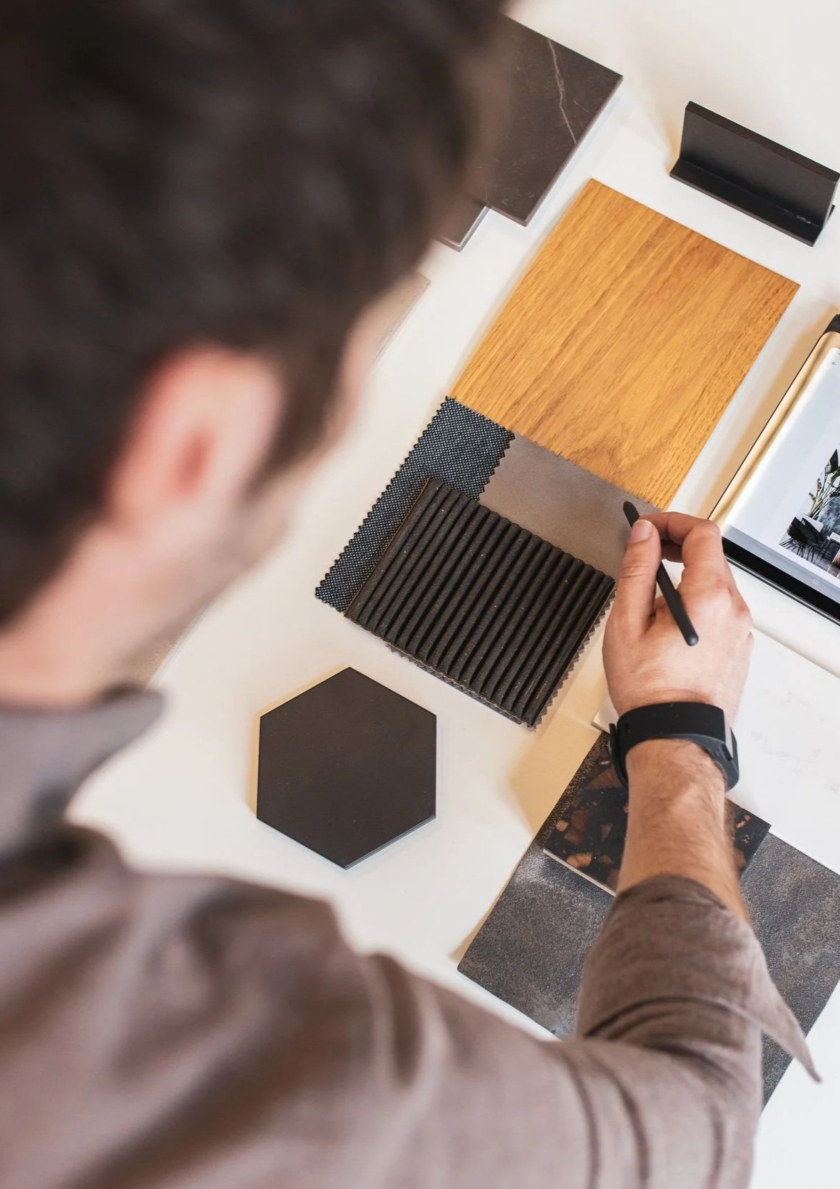 A person is designing or selecting materials for interior decoration, observing various material samples on a table, including wood, fabric, tile, metal, and stone patterns.