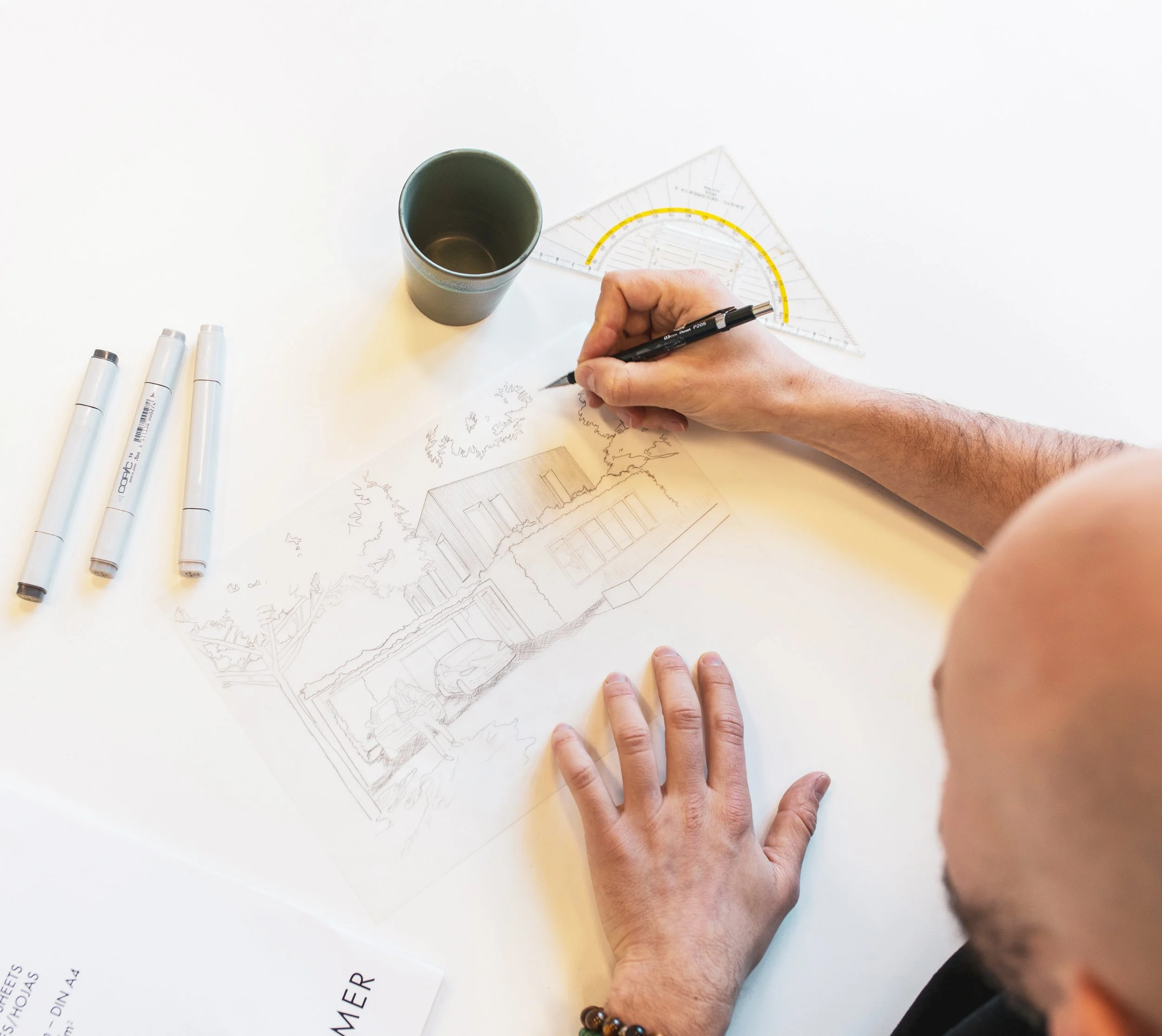 A person drawing architectural plans at a white table with markers and a transparent triangle ruler, a black coffee mug, and a white paper with text on it.