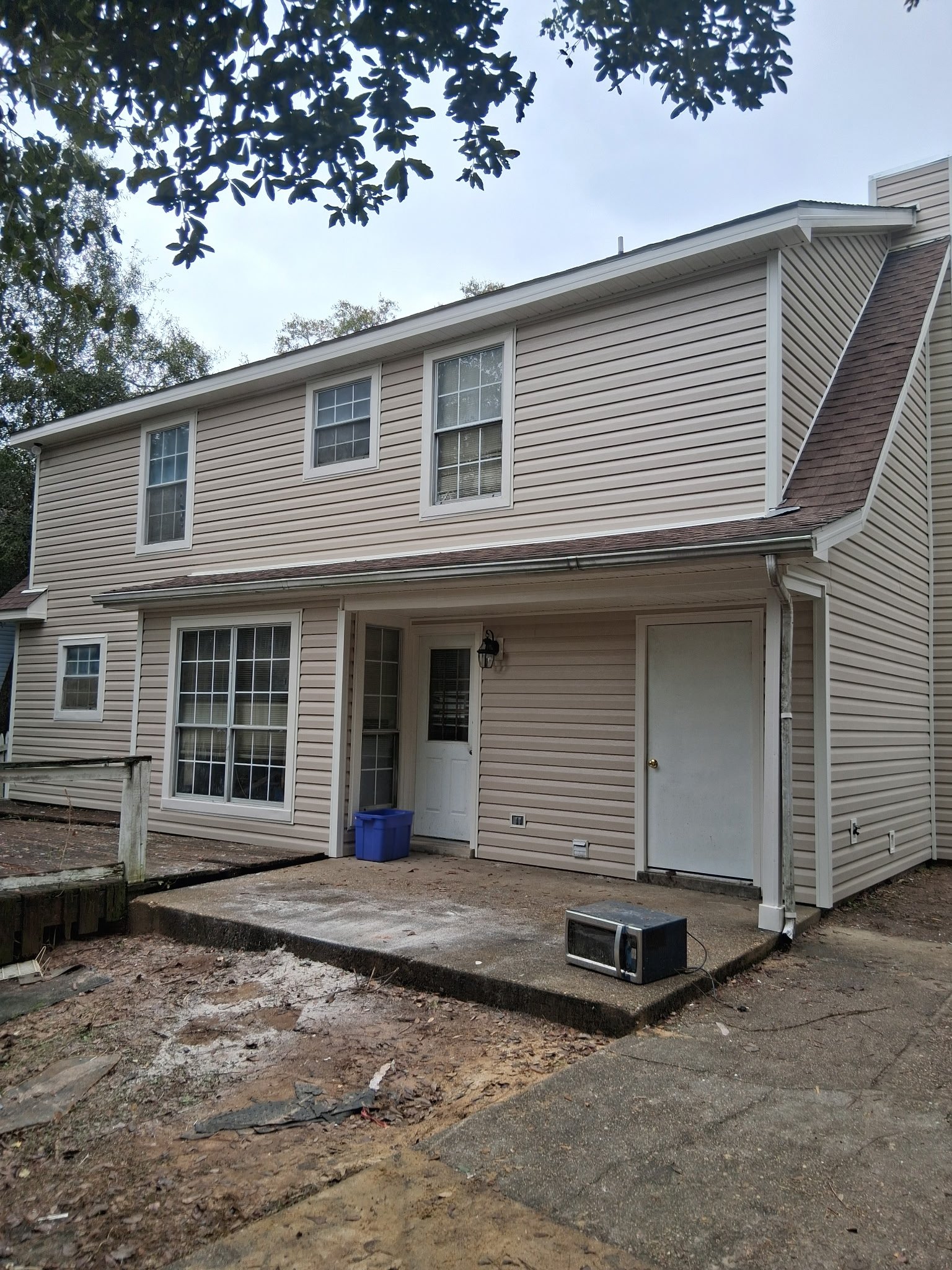 A two-story house with beige vinyl siding, white trim, and several windows. The house has a small covered porch area with a white door and a lantern-style light. In front, there is a concrete and dirt area with some construction or yard work debris, a blue recycling bin, and an old black television on the ground. The sky is overcast, and a tree branch with leaves is visible at the top of the image.
