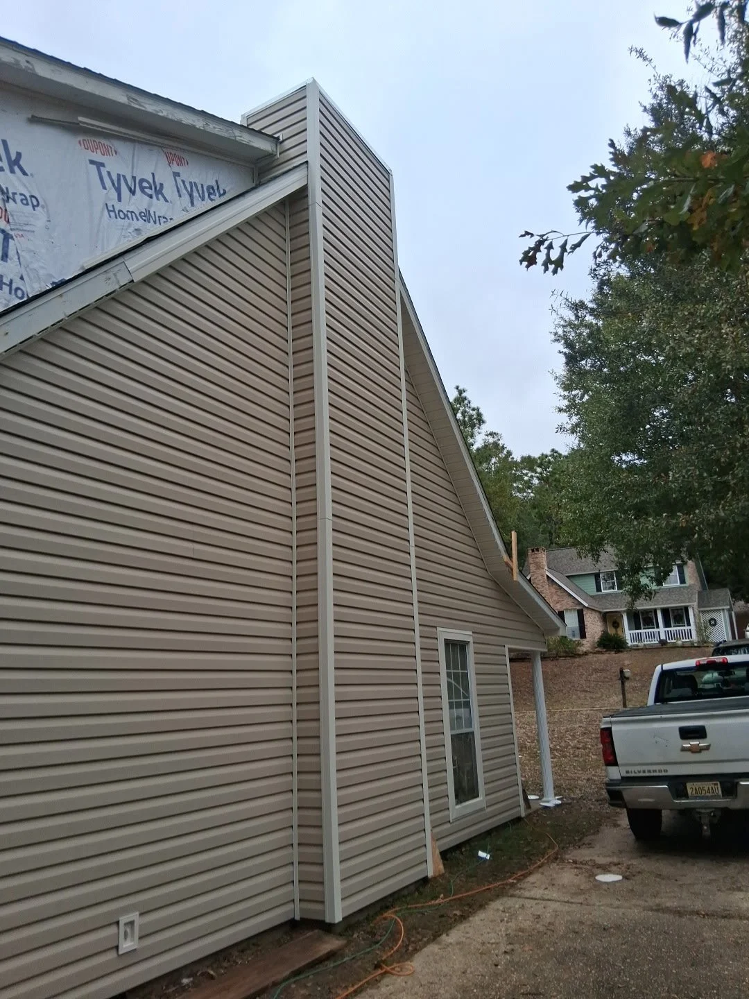 Side view of a house under construction with beige vinyl siding, a window, a white downspout, and a truck parked in the driveway, with neighboring houses and trees in the background.
