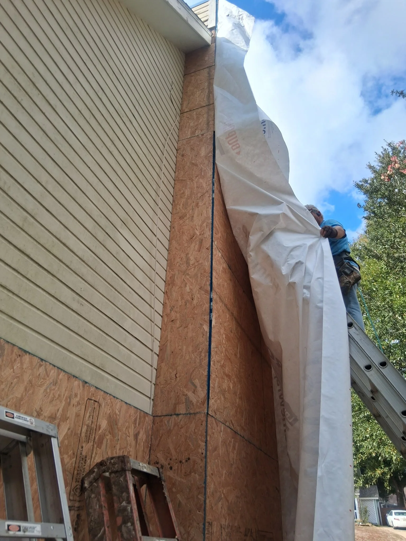 Construction worker installing a siding panel on the exterior wall of a building. The wall is partially covered with oriented strand board (OSB) sheathing, and a ladder is visible in the lower left corner. The sky is blue with some clouds, and trees are visible in the background.