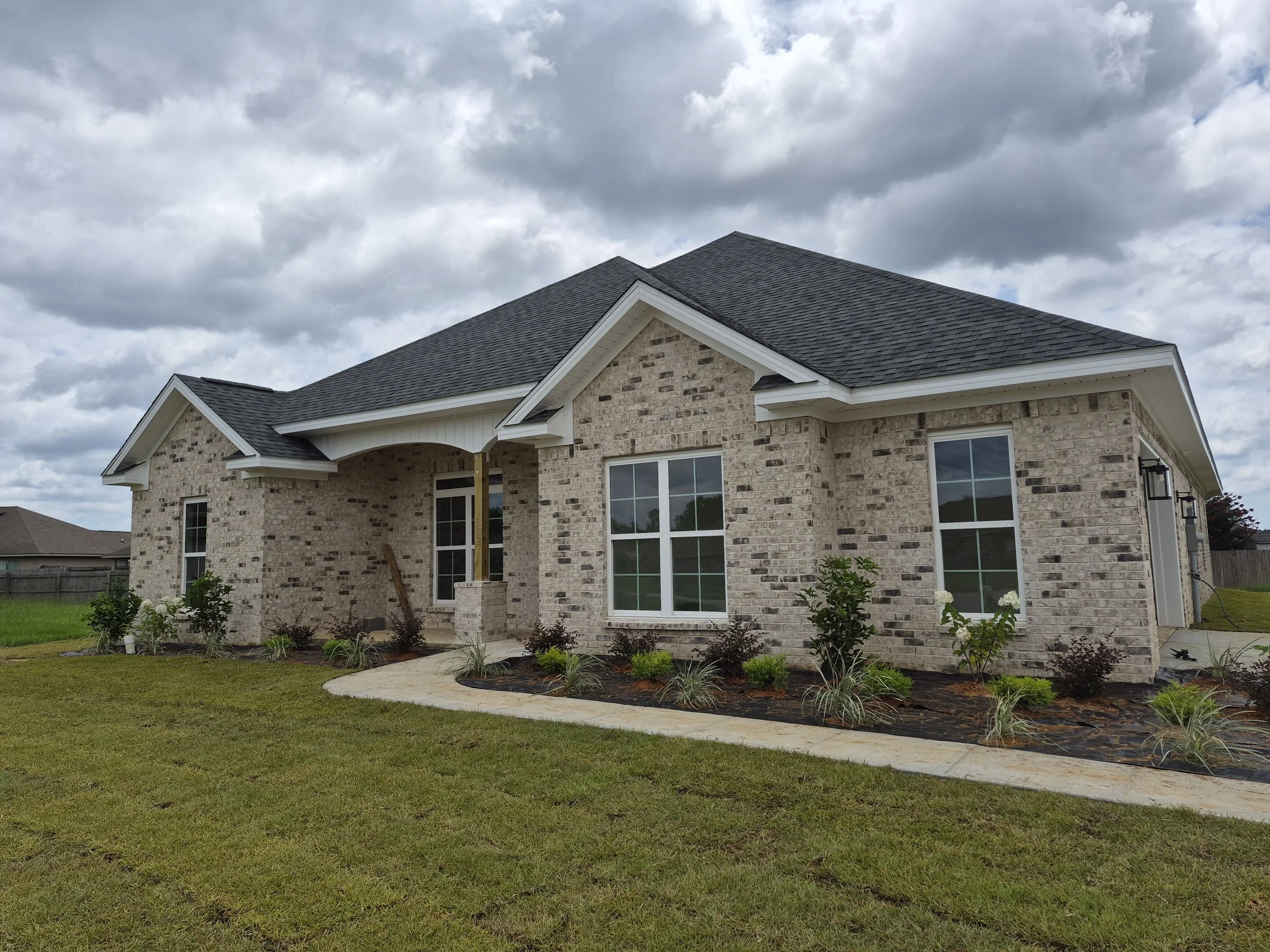 A house with brick exterior, white trim, and large windows, set against a cloudy sky. There is a porch, a garden with plants, and a grassy lawn in the front yard.