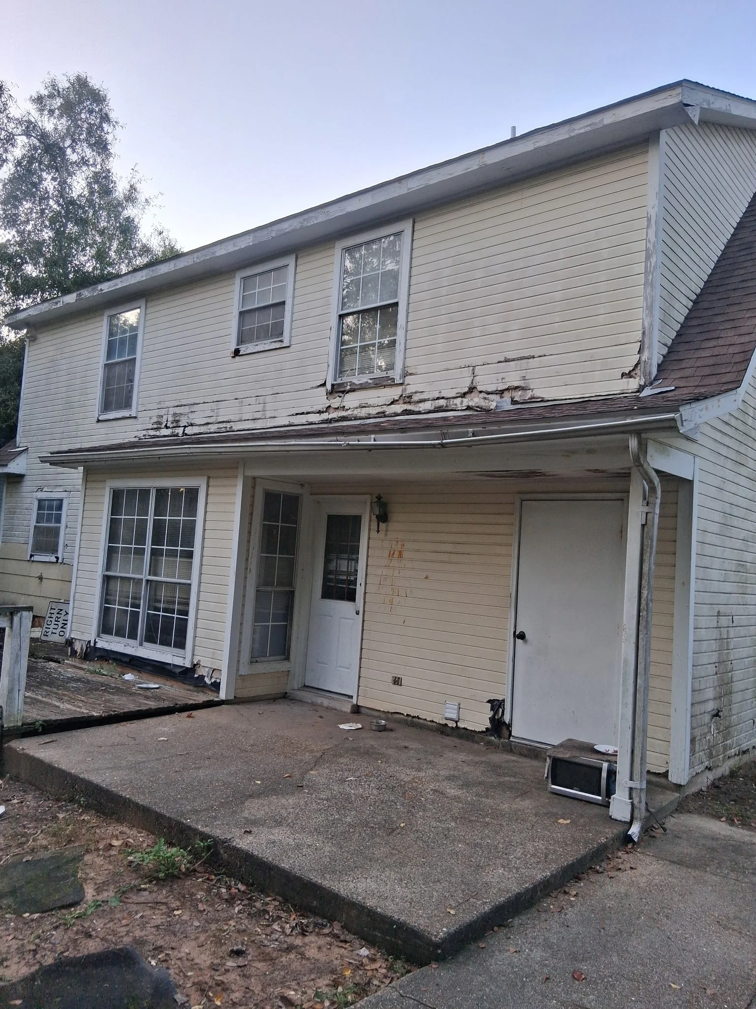 The front of a two-story house with peeling paint and damaged siding, with a concrete porch and fallen debris, including an old microwave on the ground.