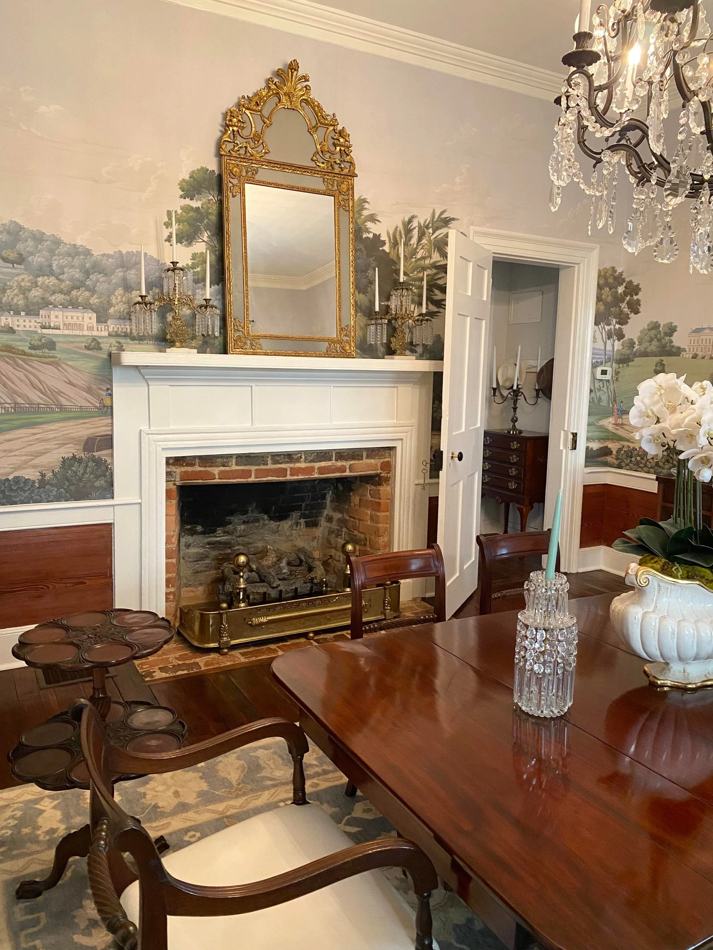 A dining room featuring a large wooden table, upholstered chairs, a chandelier, a fireplace with a mirror, wall sconces, and a mural wall painting of a landscape with trees, hills, and buildings.