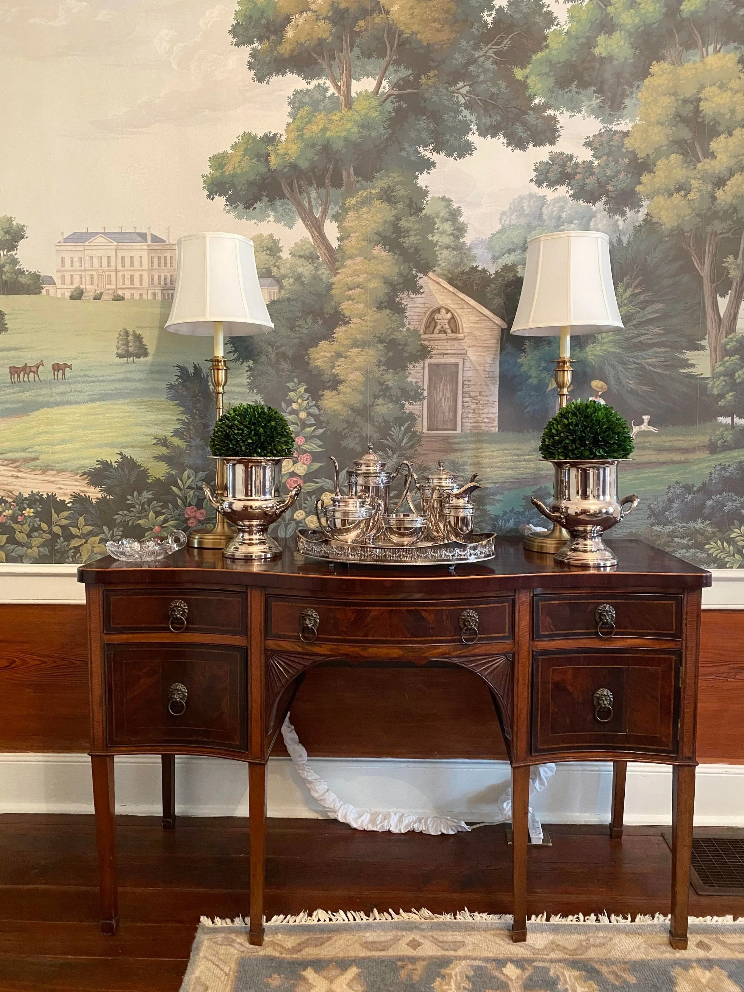A wooden sideboard with decorative metal handles, topped with two table lamps with white shades, two small topiary plants, and a silver tea set. In the background, there is a large mural of a scenic landscape featuring trees, a small stone building, a grand mansion, horses grazing, and a woman with a parasol.
