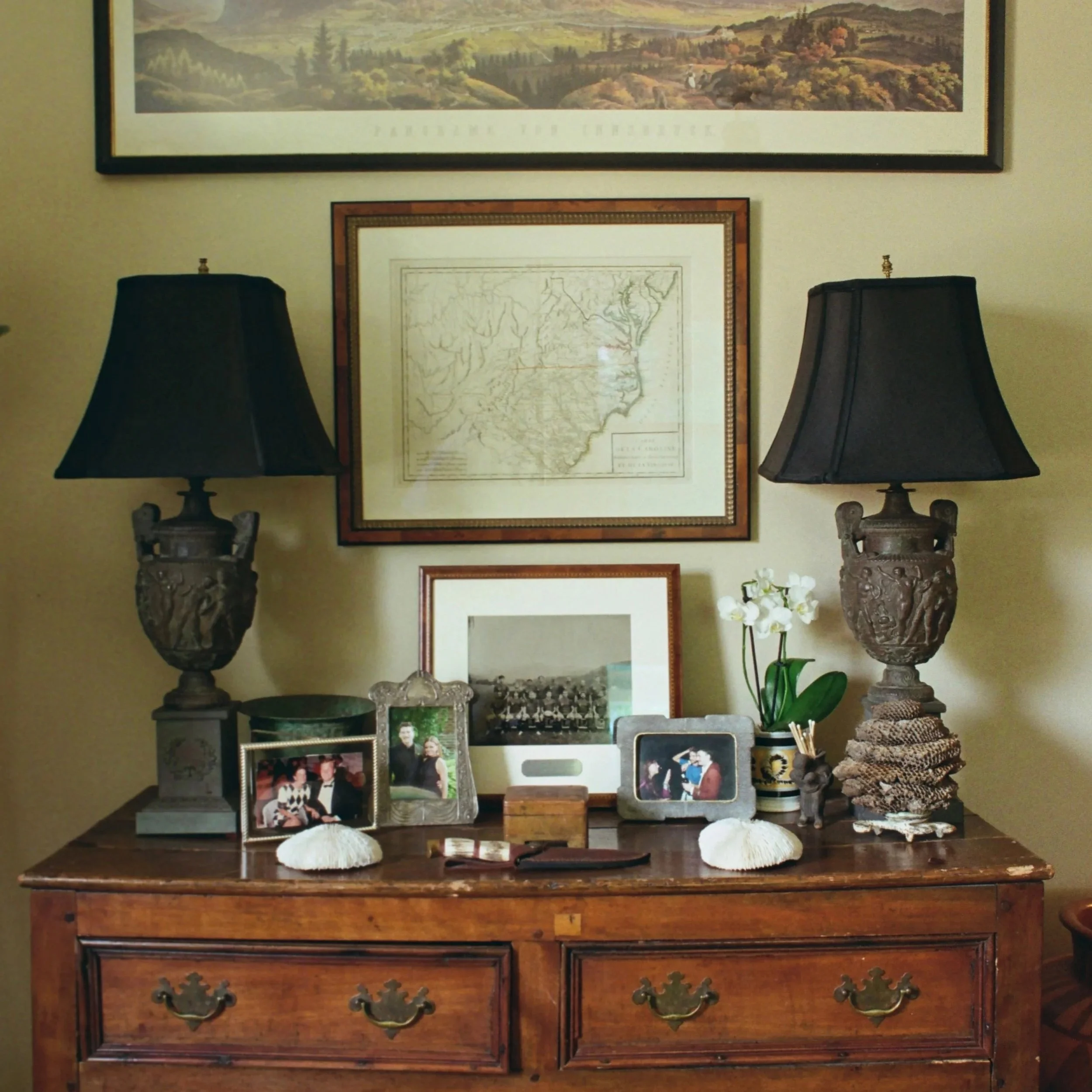 A wooden sideboard with two large black lamps, framed pictures including a map and photographs, decorative shells, flowers, and other small decor items on top, with a large landscape painting hanging above.