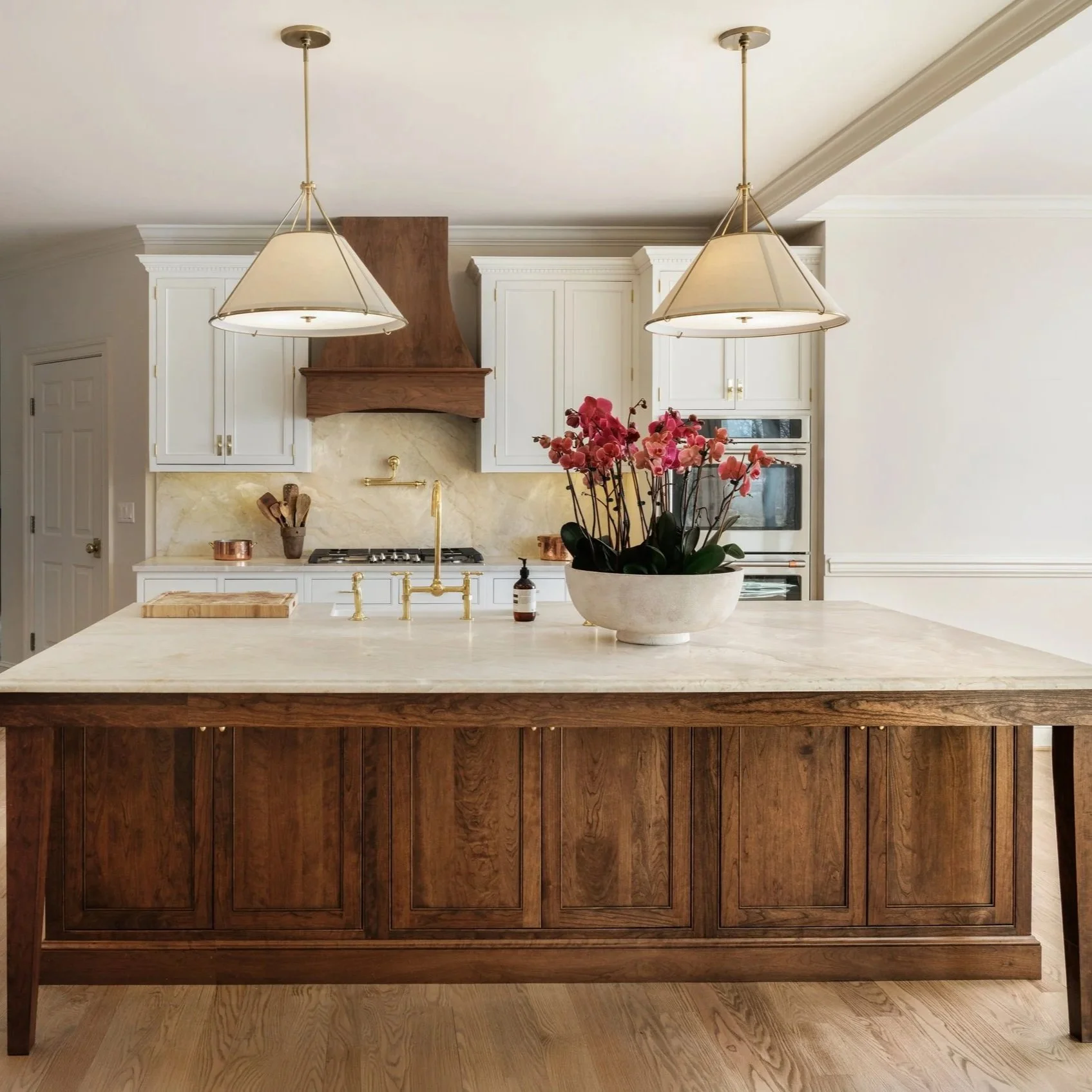 A kitchen island with a white marble top, brass faucet, and a potted orchid plant. In the background, there are white cabinets, a wooden range hood, and a built-in oven. Two pendant lights hang from the ceiling.