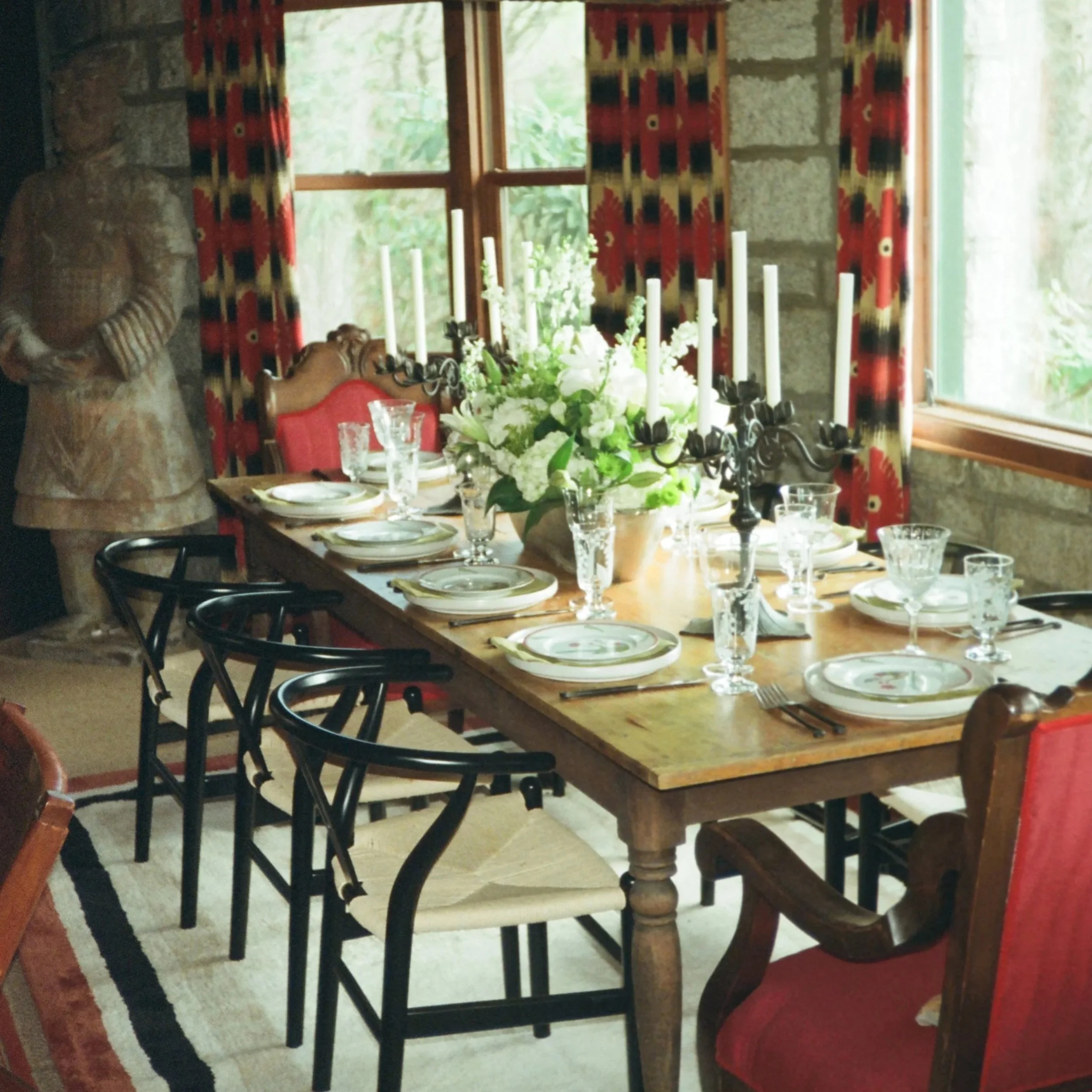 A dining room with a long wooden table set with plates, glasses, and a centerpiece of white and green flowers. Black chairs surround the table. Behind the table, there is a window with floral curtains, and a stone wall is visible on the left. A manne